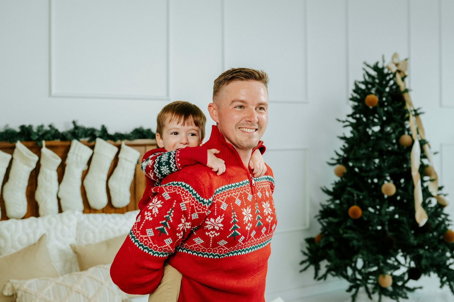 Two people wearing matching red holiday sweaters pose in front of a decorated Christmas tree and stockings.