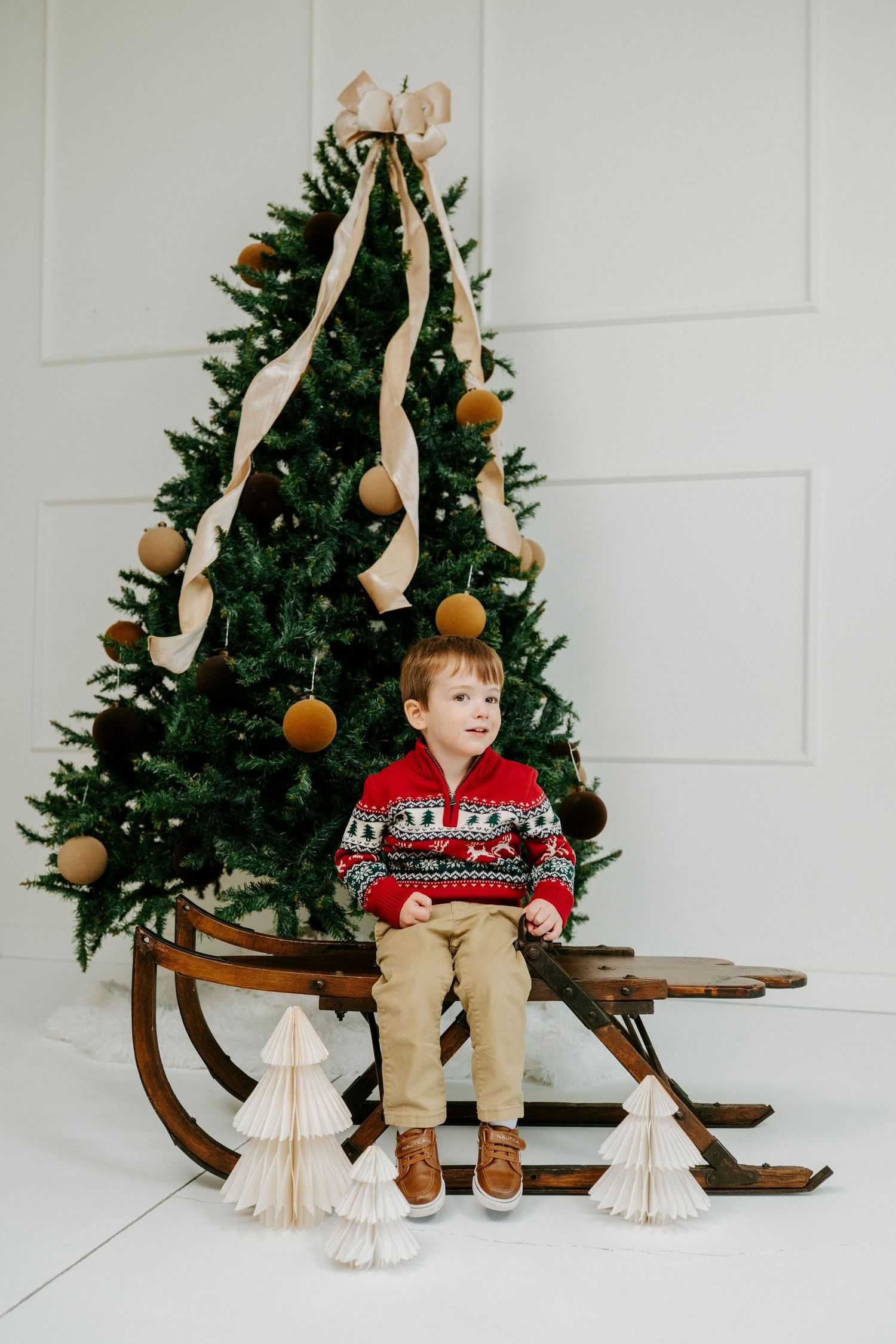 A vintage wooden sled sits in front of a Christmas tree decorated with gold ornaments and cream ribbons.