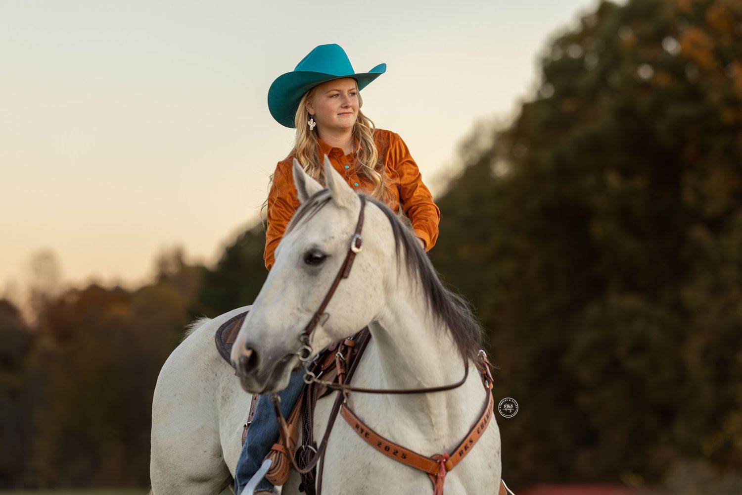 White horse and rider stand in a fall meadow with vibrant orange autumn foliage in the background.