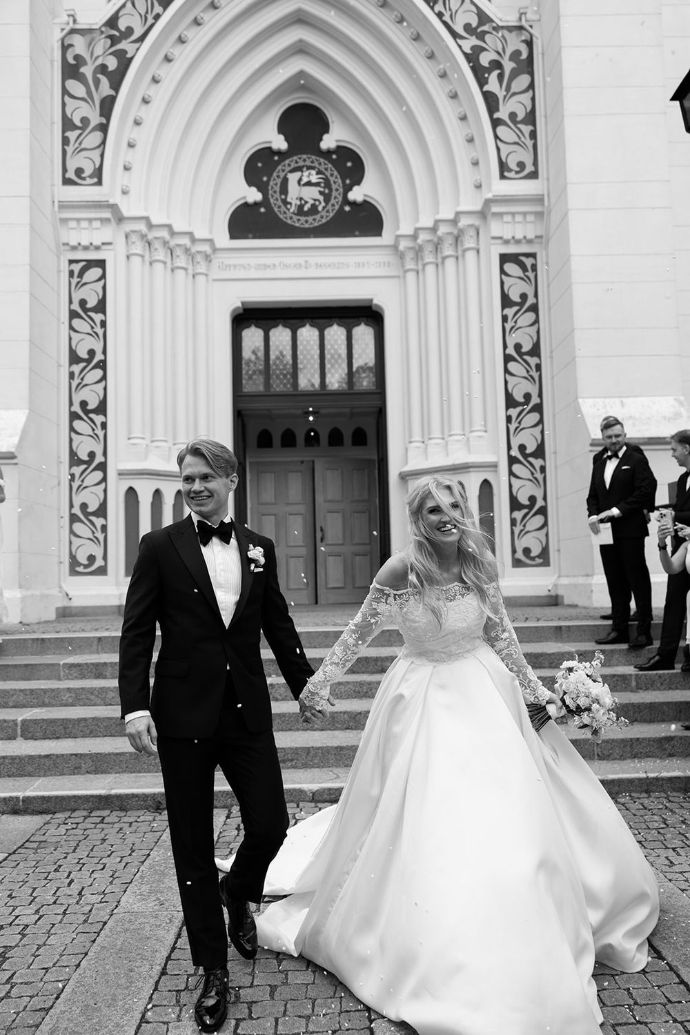 Bröllopsfotograf Emelie Petré, A wedding couple walks outside a grand white church with ornate arched doorways and stone facade.
