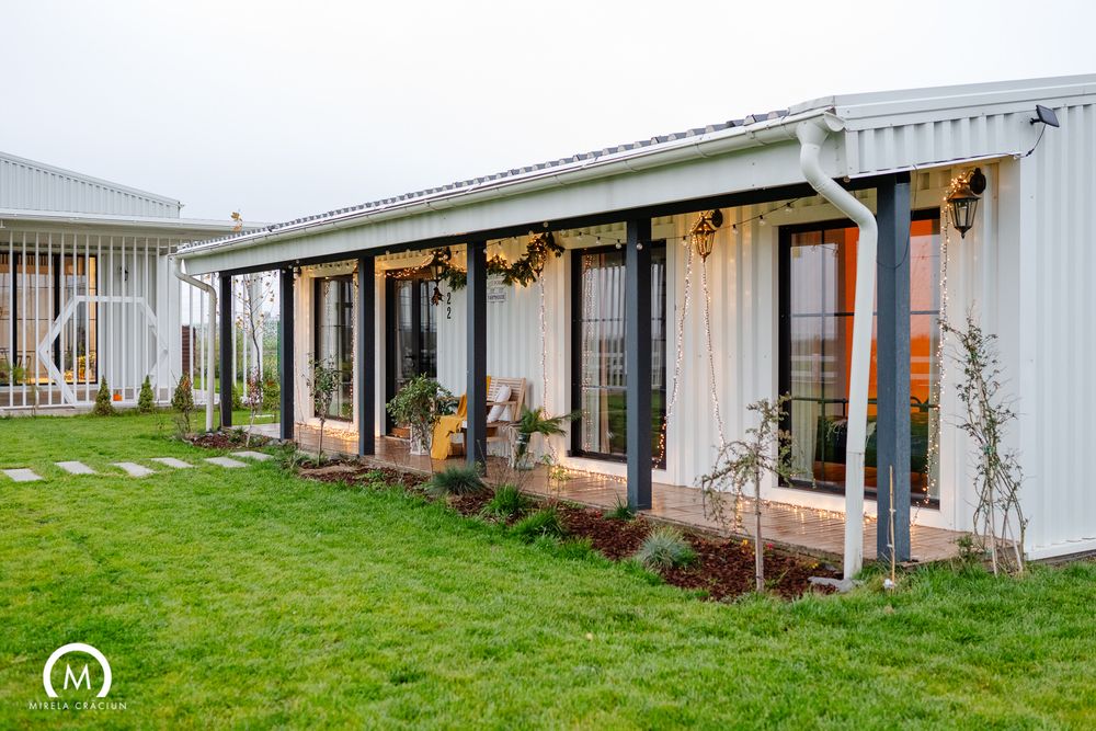 Modern shipping container home with rustic porch decorated with autumn garlands and pumpkins on green lawn.