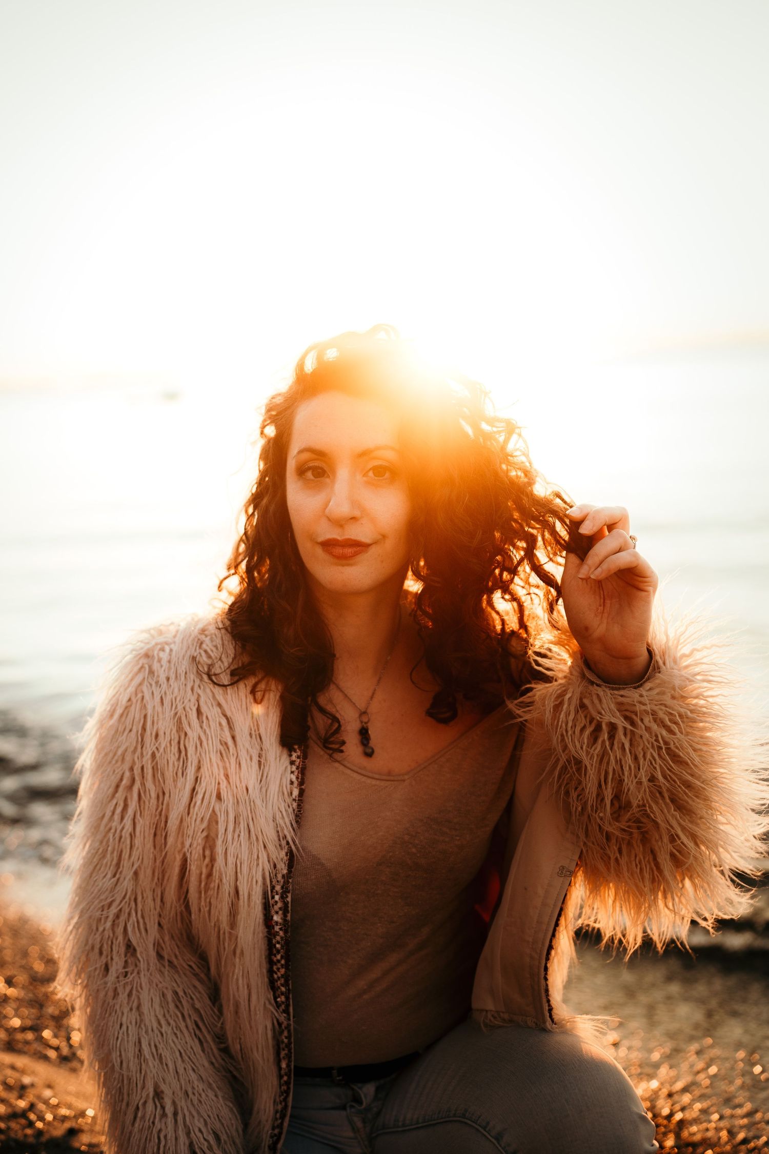 A person in a fur coat poses on a beach at sunset with warm golden light creating a dreamy atmosphere.