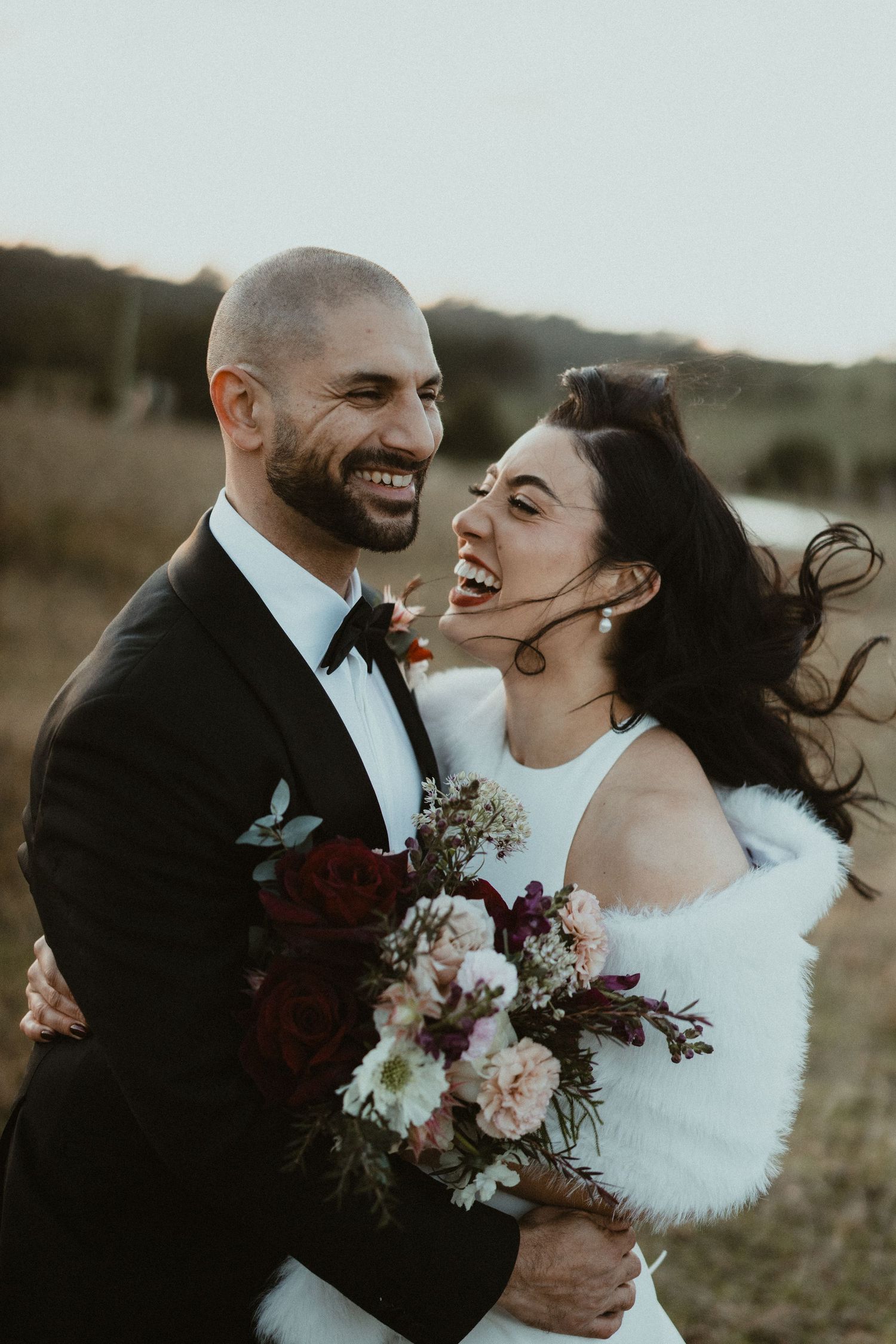 Joyful wedding couple sharing laughter during outdoor portrait session with floral bouquet.