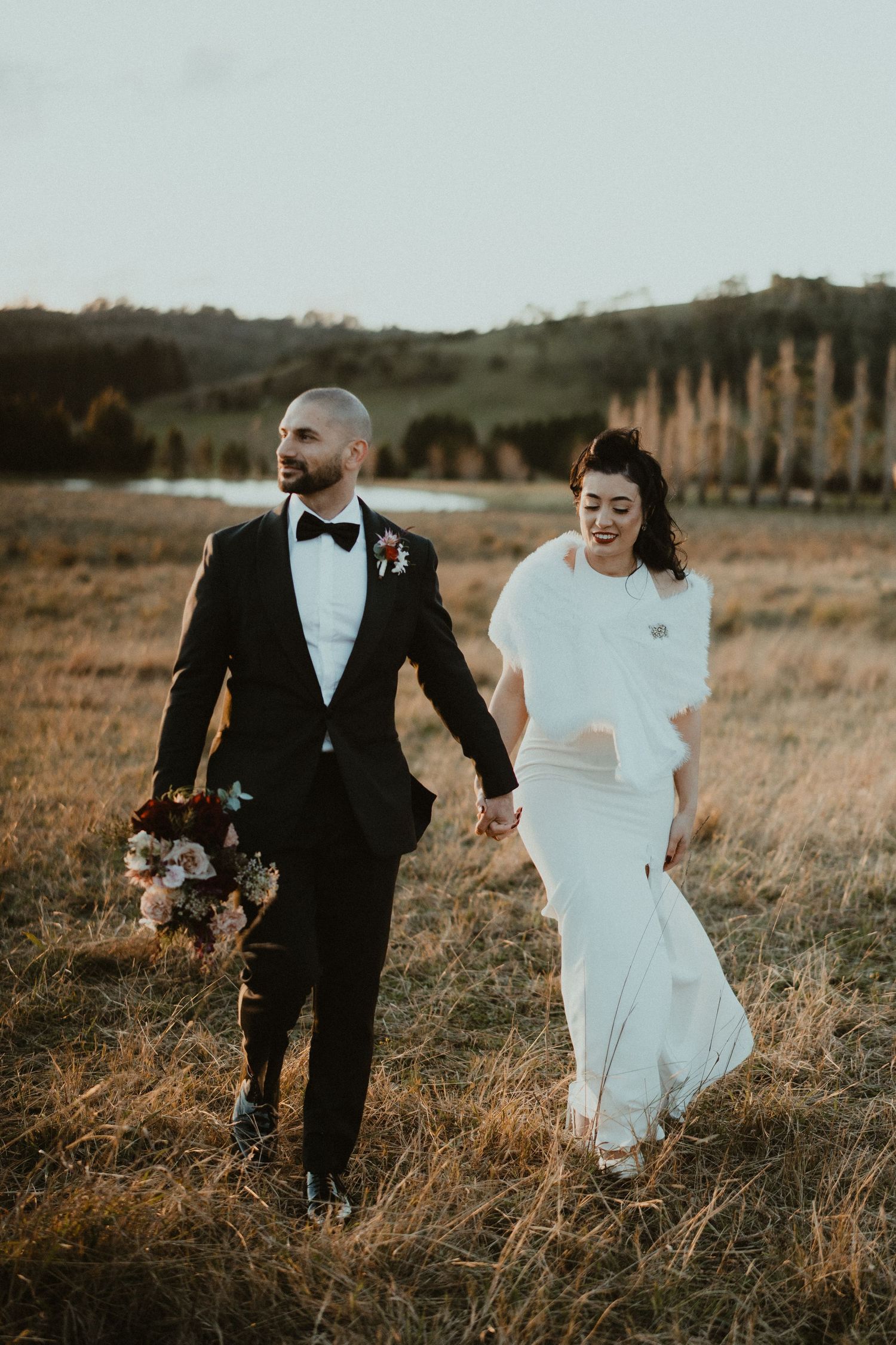 Newlyweds walking through a golden field at sunset in formal wedding attire.
