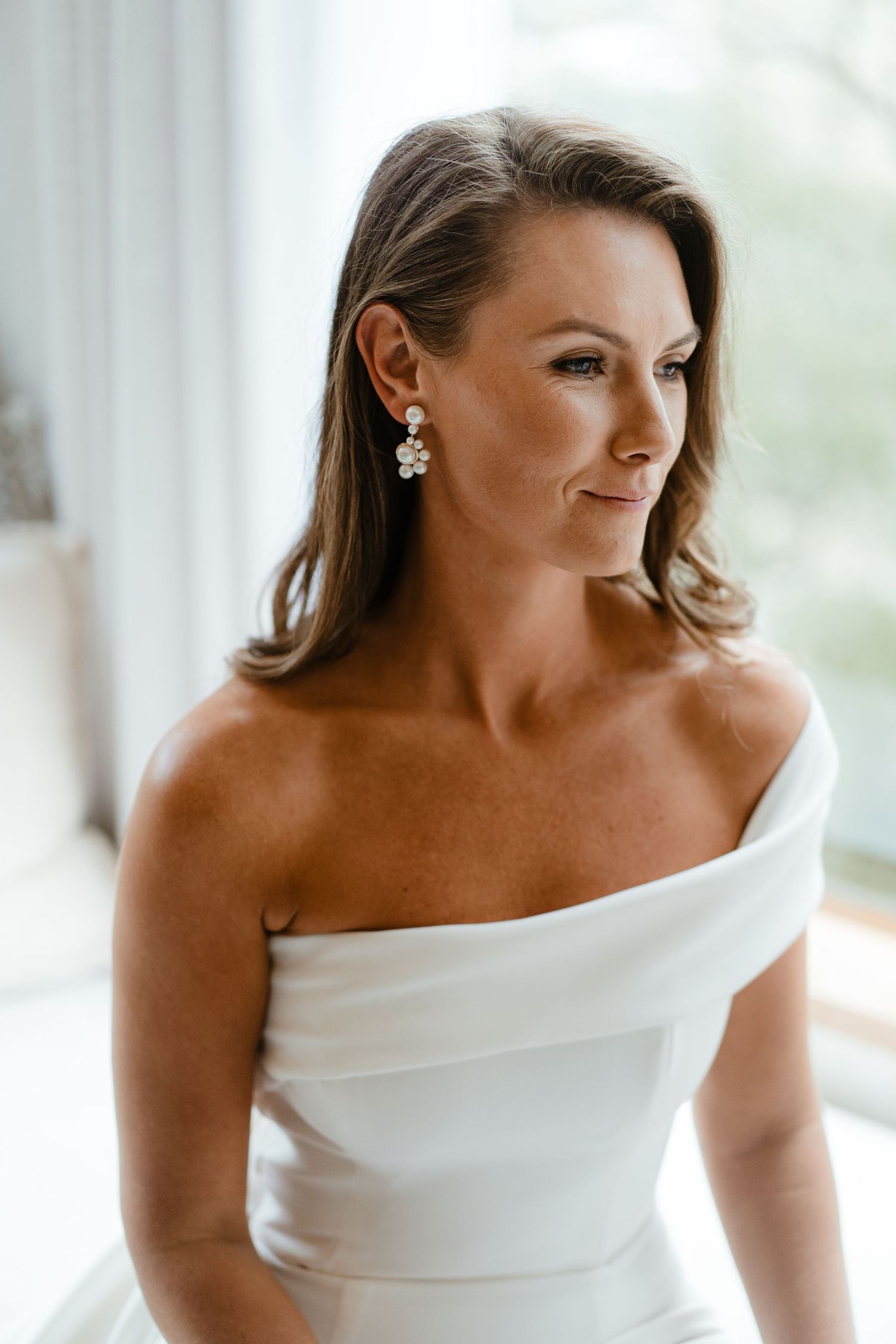Elegant bride in modern off-shoulder white wedding dress posing by window.