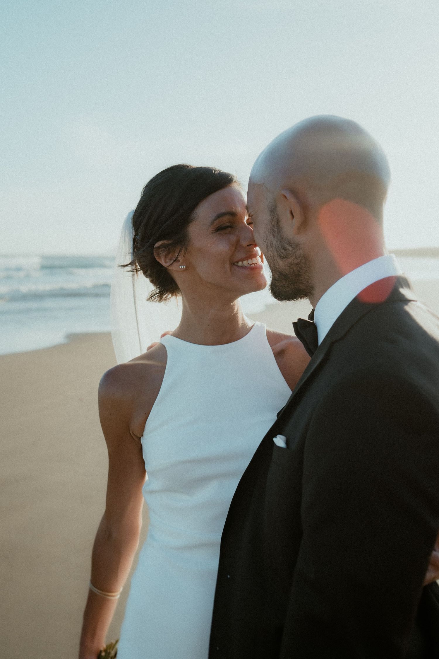Couple in wedding attire sharing intimate moments against a soft, bright coastal background.