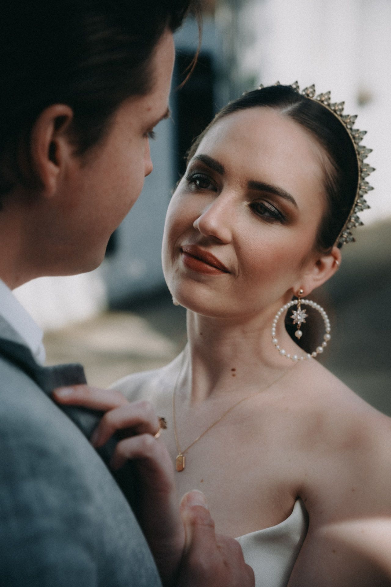 A close-up wedding portrait showing a bride wearing a delicate crown and statement earrings gazing lovingly at her partner.