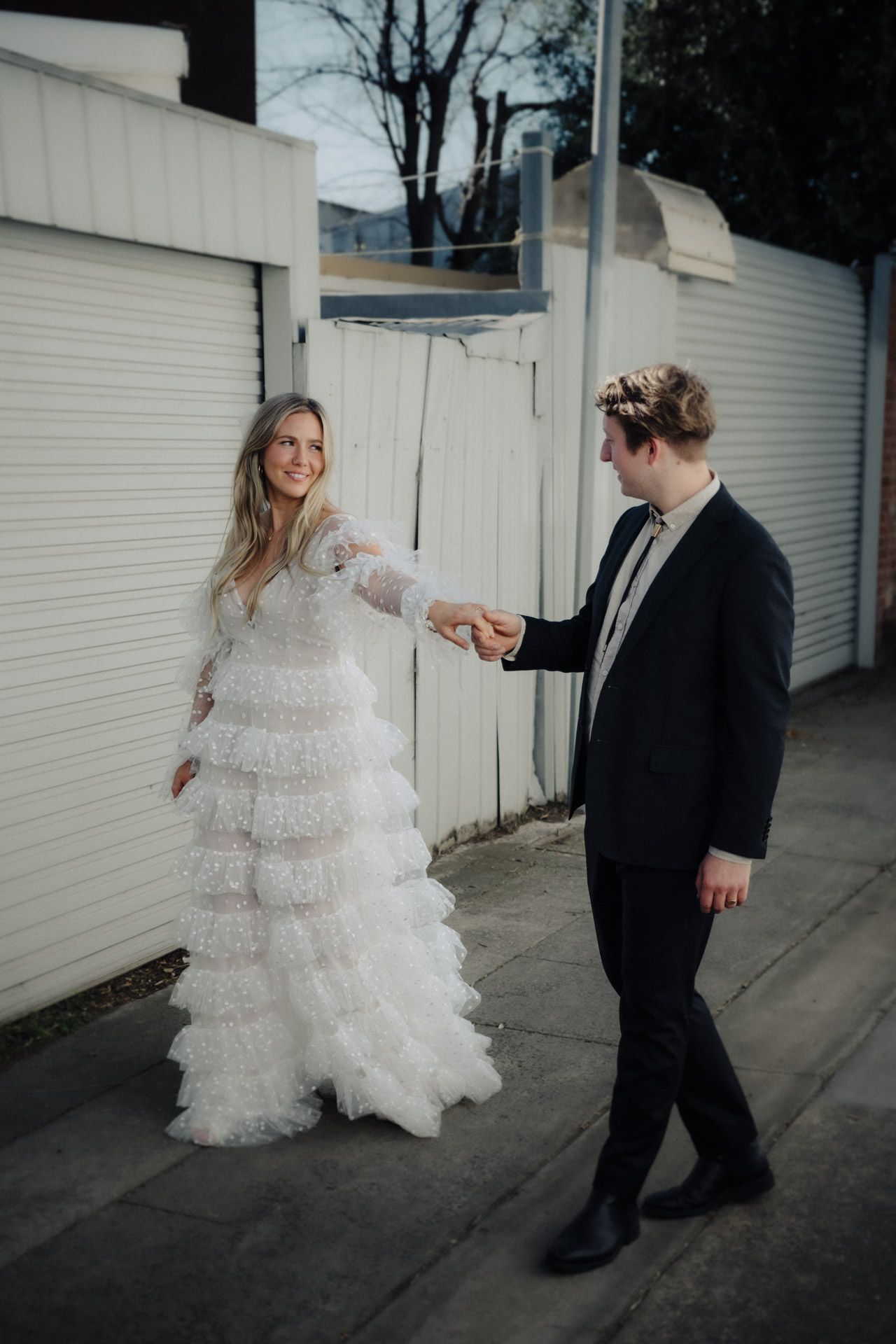 Couple dances playfully outside white storage units in dramatic wedding attire.