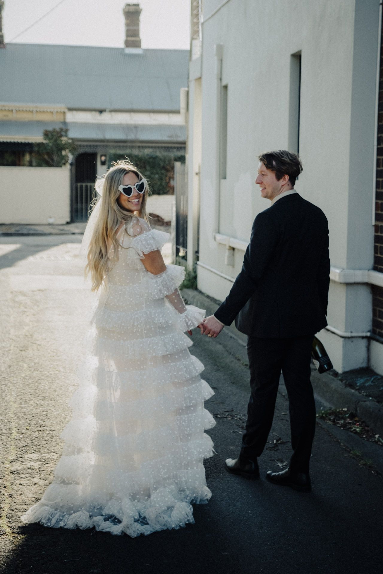 Couple in wedding attire walking together on outdoor pathway with sunlit backdrop.