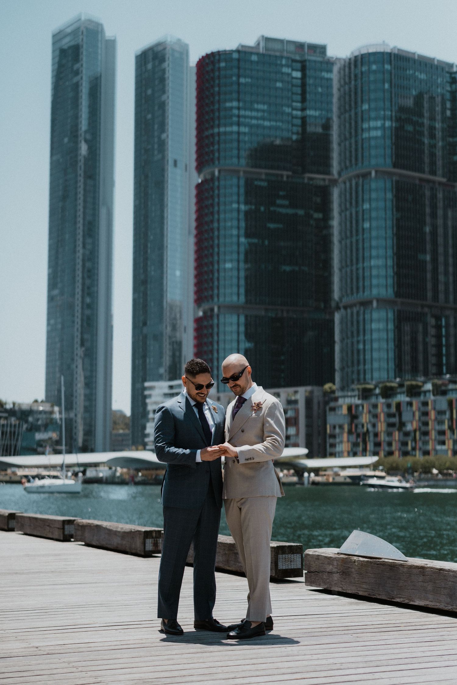 Two figures in business attire converse on a wooden boardwalk with modern glass buildings and a harbor in the background.