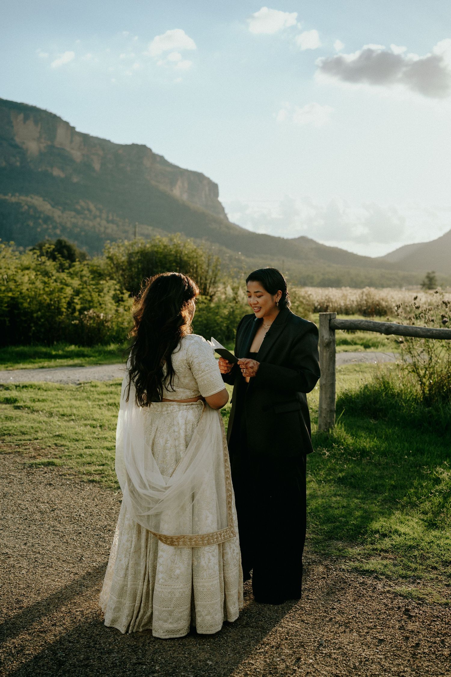 Two figures in contrasting white and black outfits embrace on a path with dramatic mountain scenery in the background.