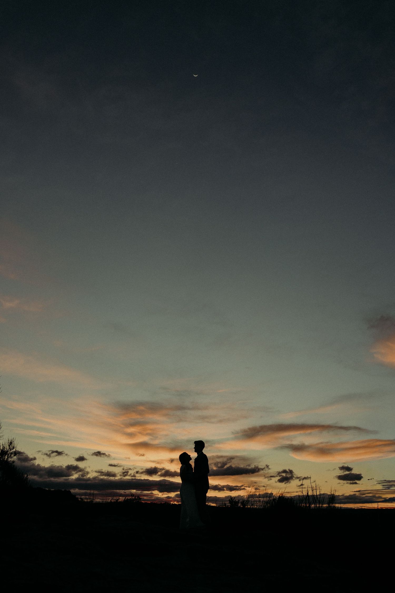 Silhouette of a couple standing together at sunset with dramatic clouds and a crescent moon in the twilight sky.