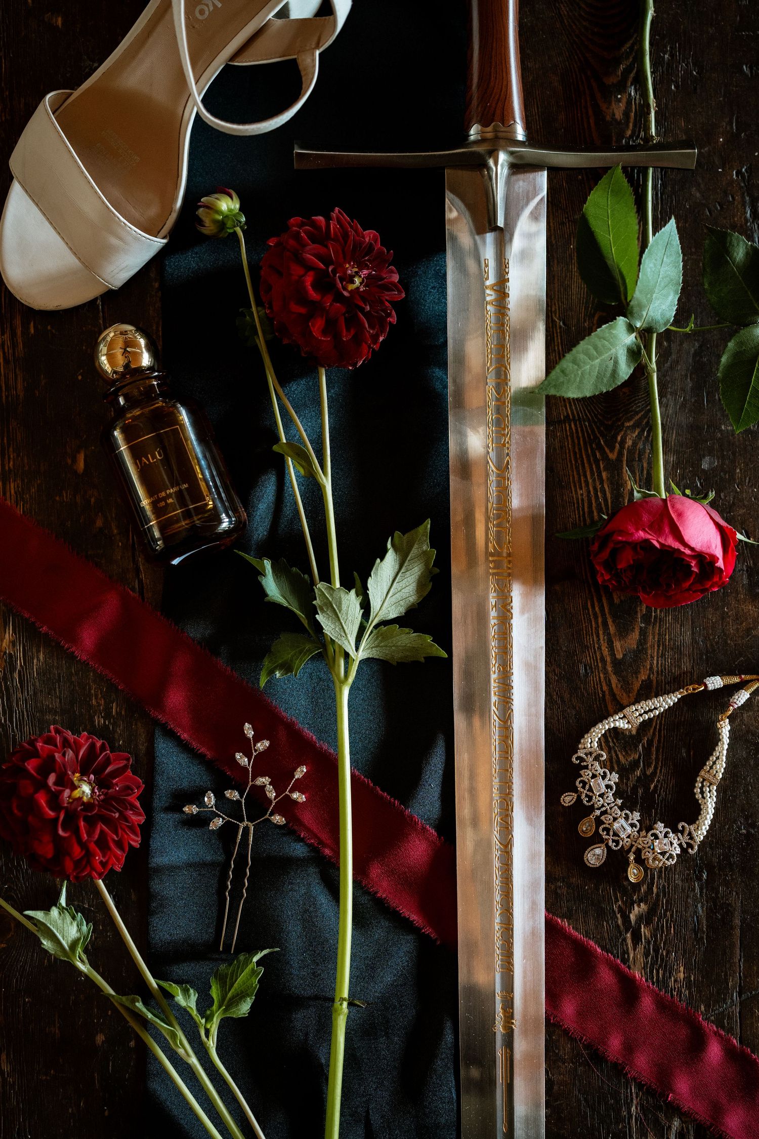 Dark romantic flatlay with sword, red dahlias, roses, pearl necklace, perfume, hair pin, red ribbon, and white heels on wood.