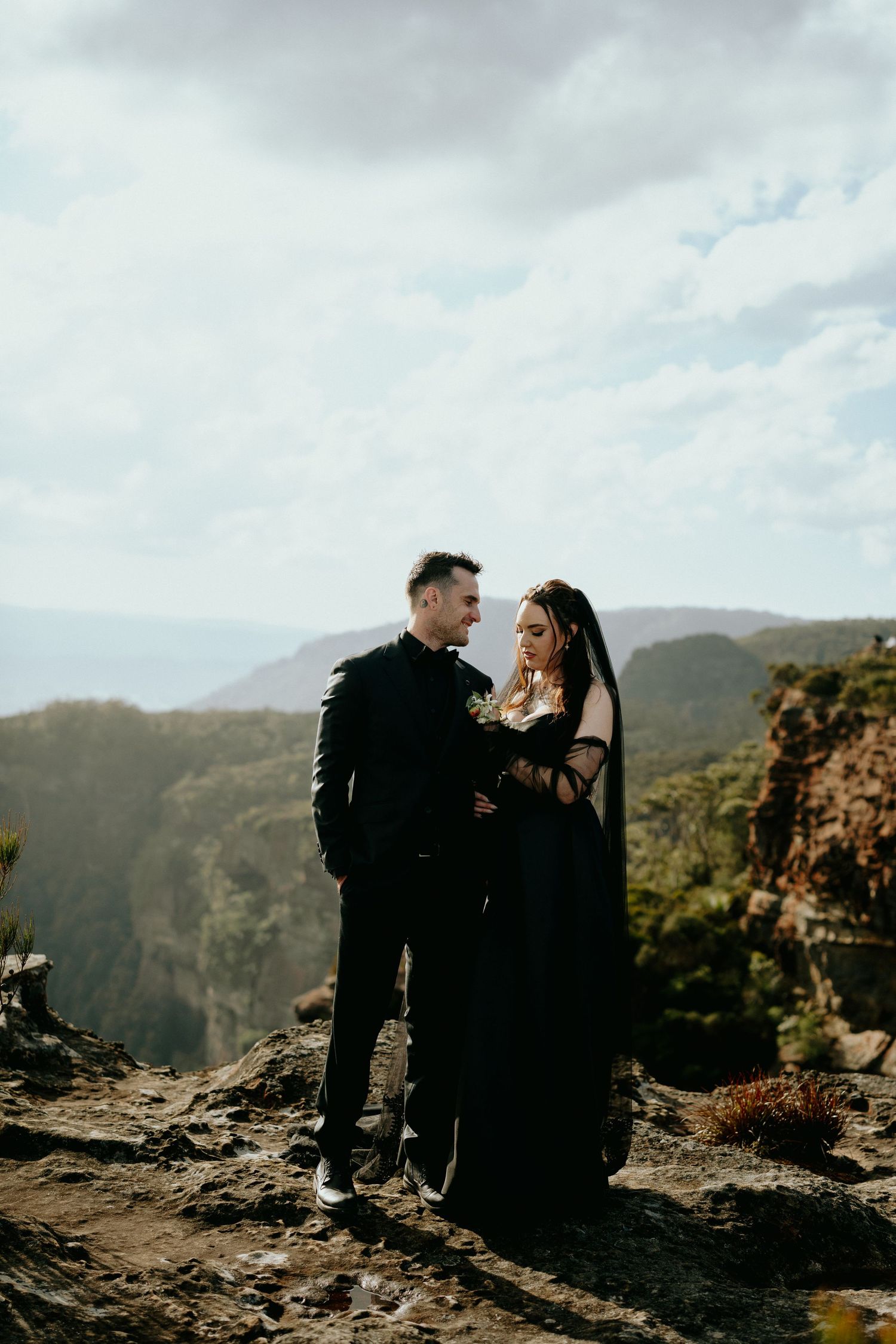 Couple stands on rocky overlook with dramatic mountain vista behind them.