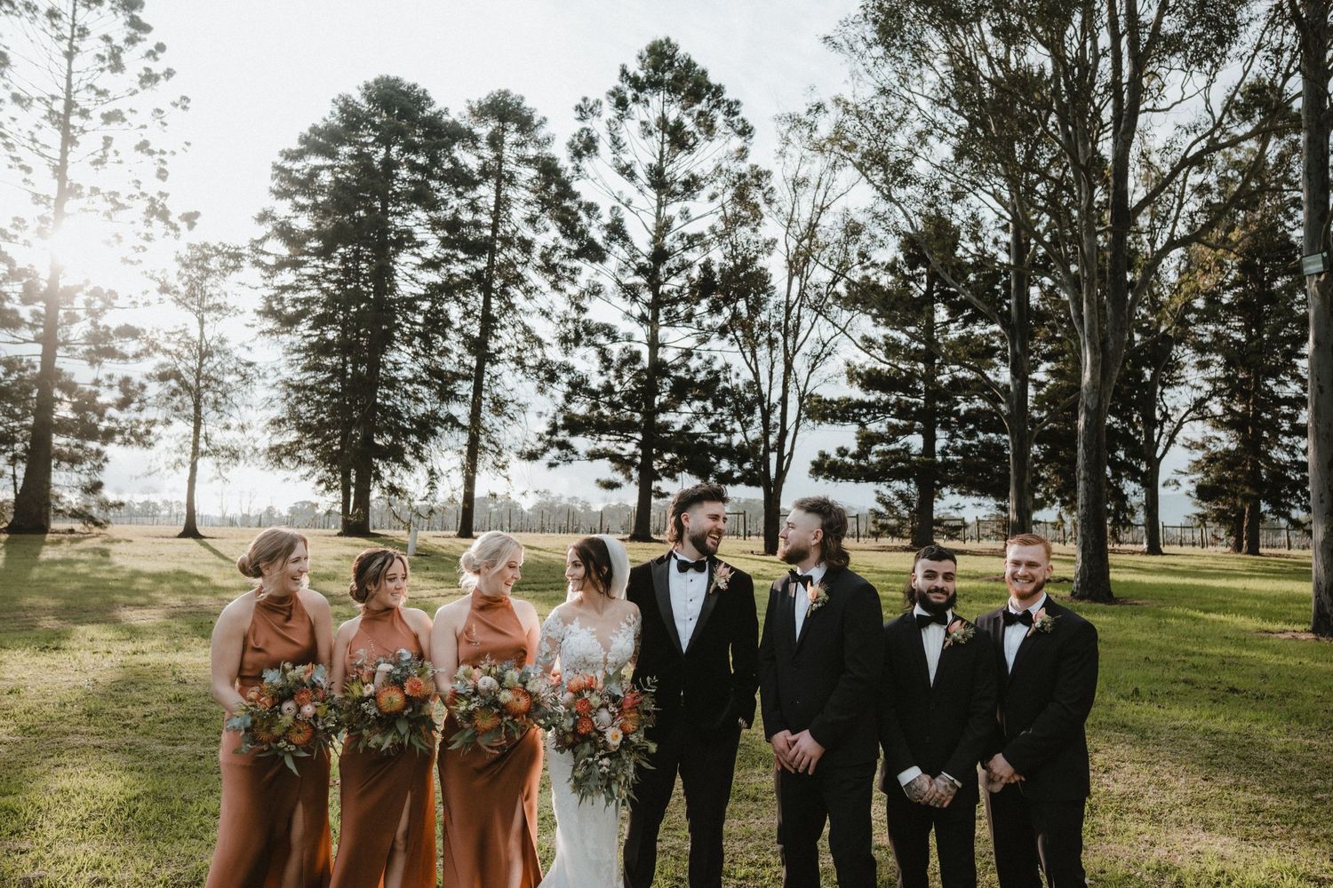Wedding party lined up outdoors with bridesmaids in rust-colored dresses and groomsmen in black suits.