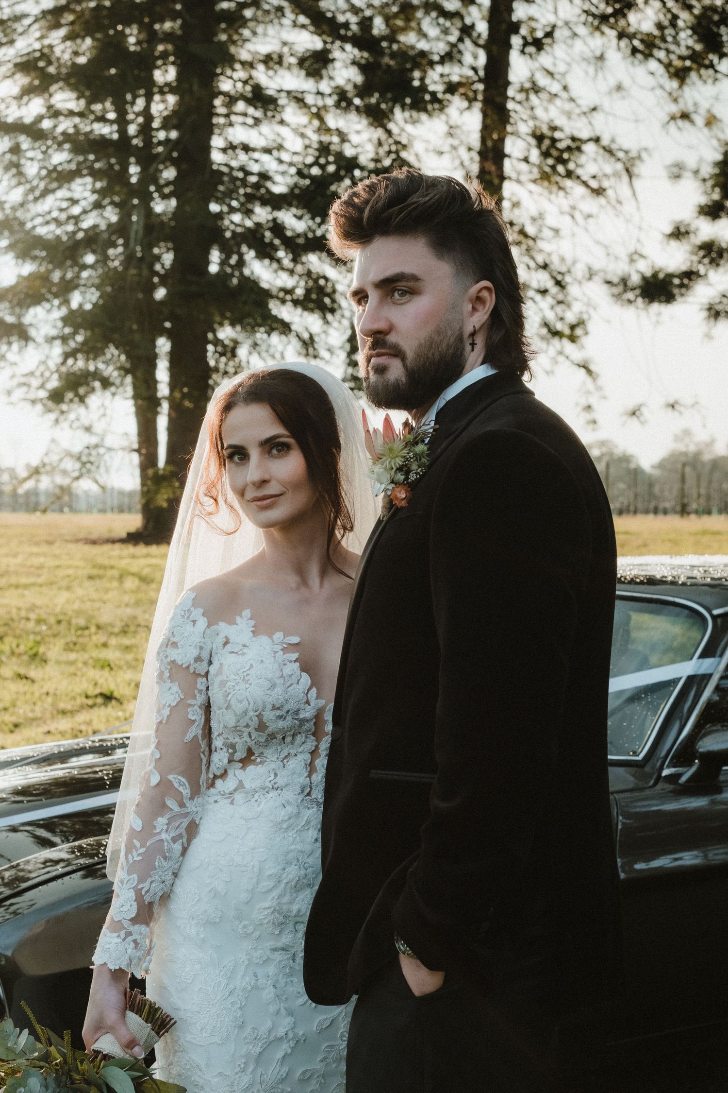 A couple poses beside a classic car during their wedding photoshoot at sunset.
