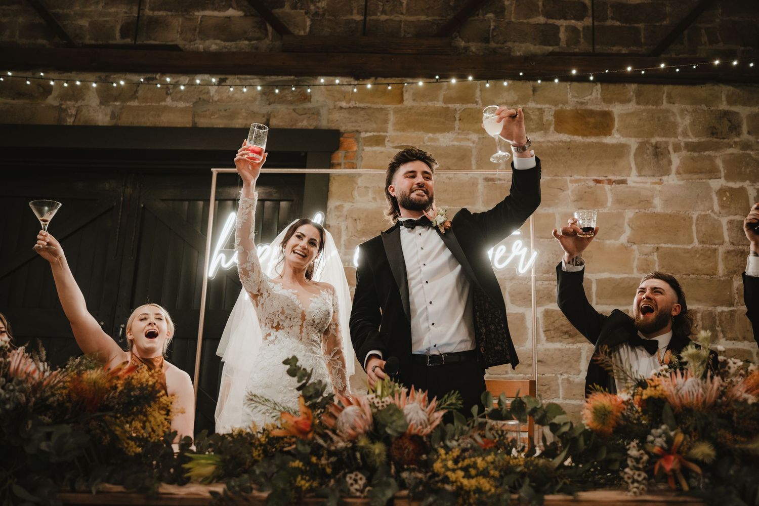 Wedding party celebrates joyfully against a rustic brick wall backdrop with string lights.