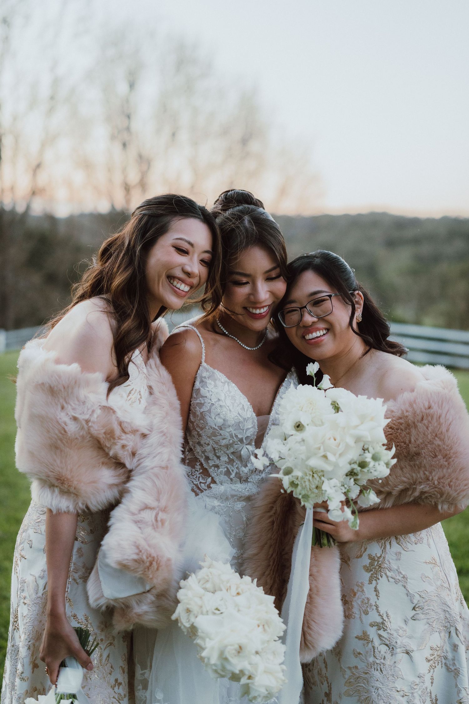 Bridesmaids in blush pink dresses share a joyful moment while holding white bouquets in an outdoor setting.
