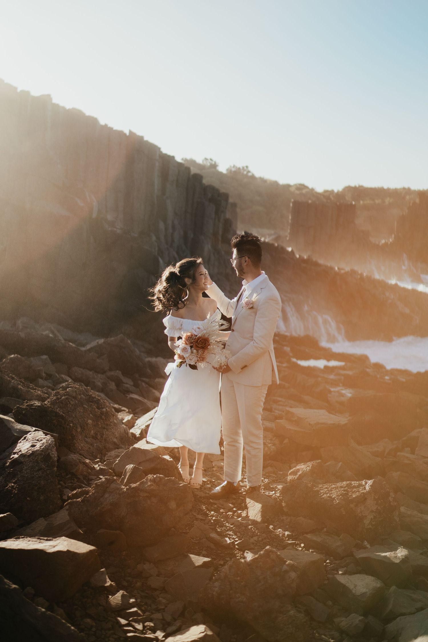 A romantic couple embracing on rocky cliffs during sunset near the ocean.