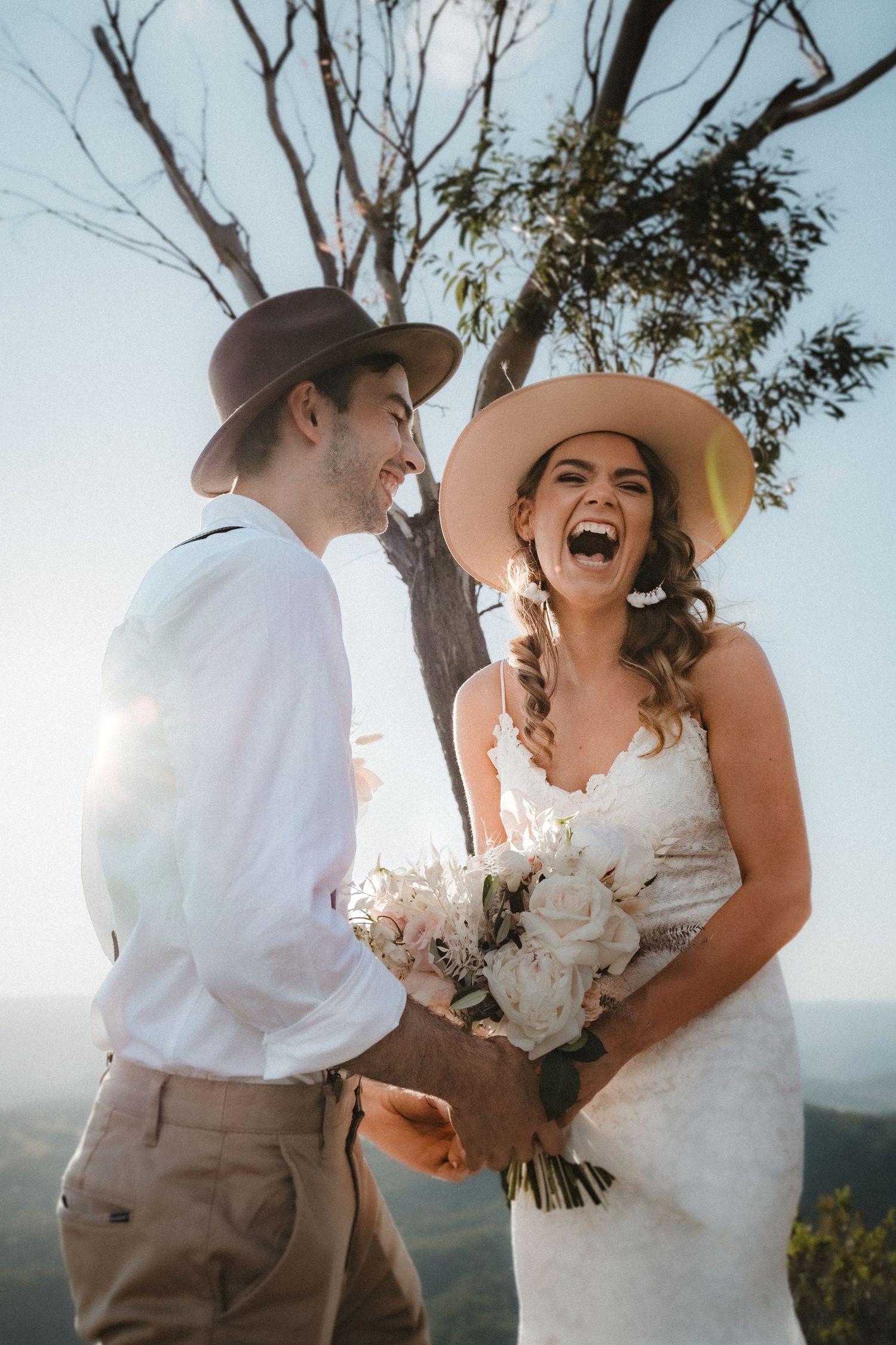 Couple wearing western hats share candid laughing moment during outdoor mountain photoshoot.