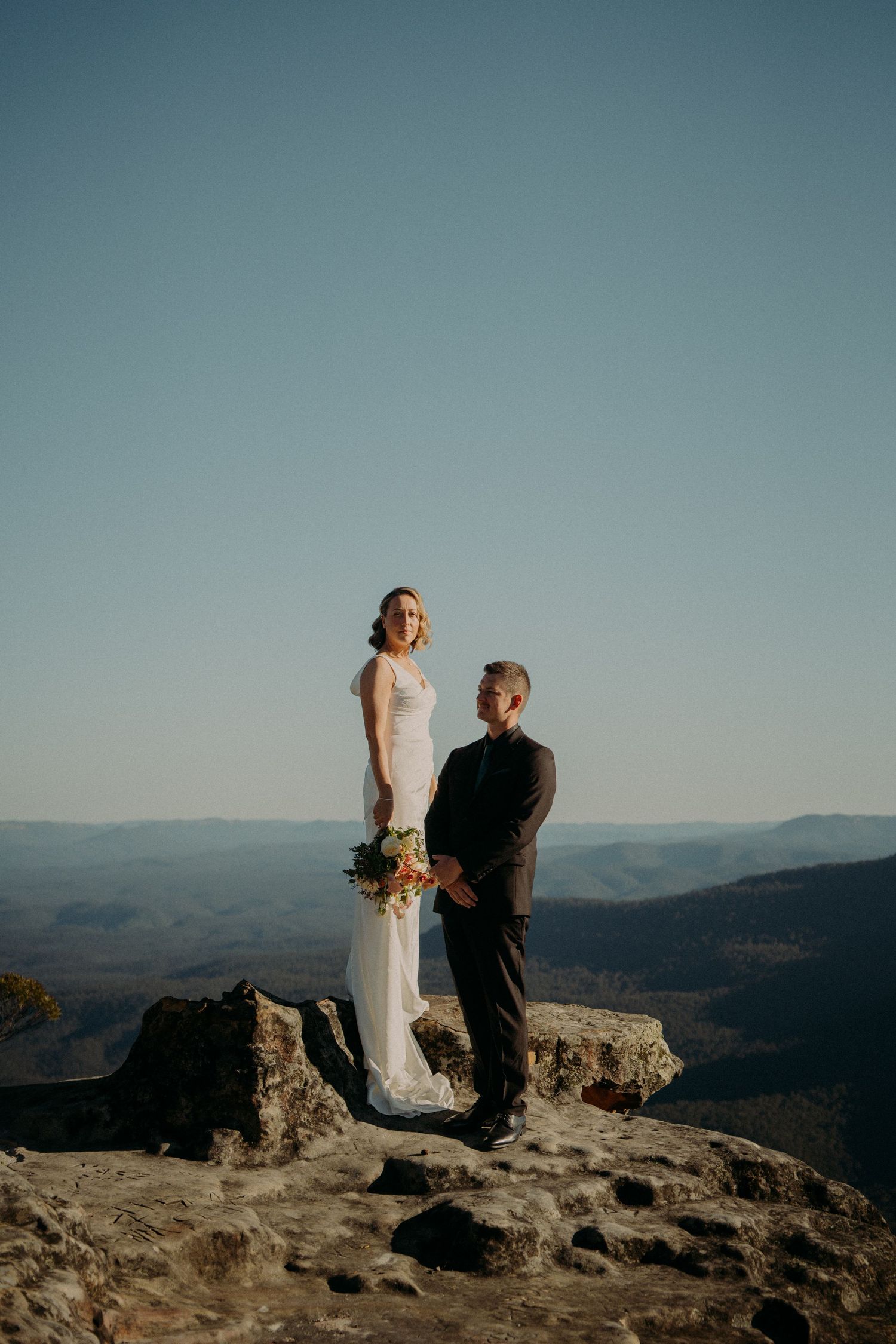Bride and groom stand on a rocky mountain summit with sweeping valley views during their outdoor elopement ceremony.