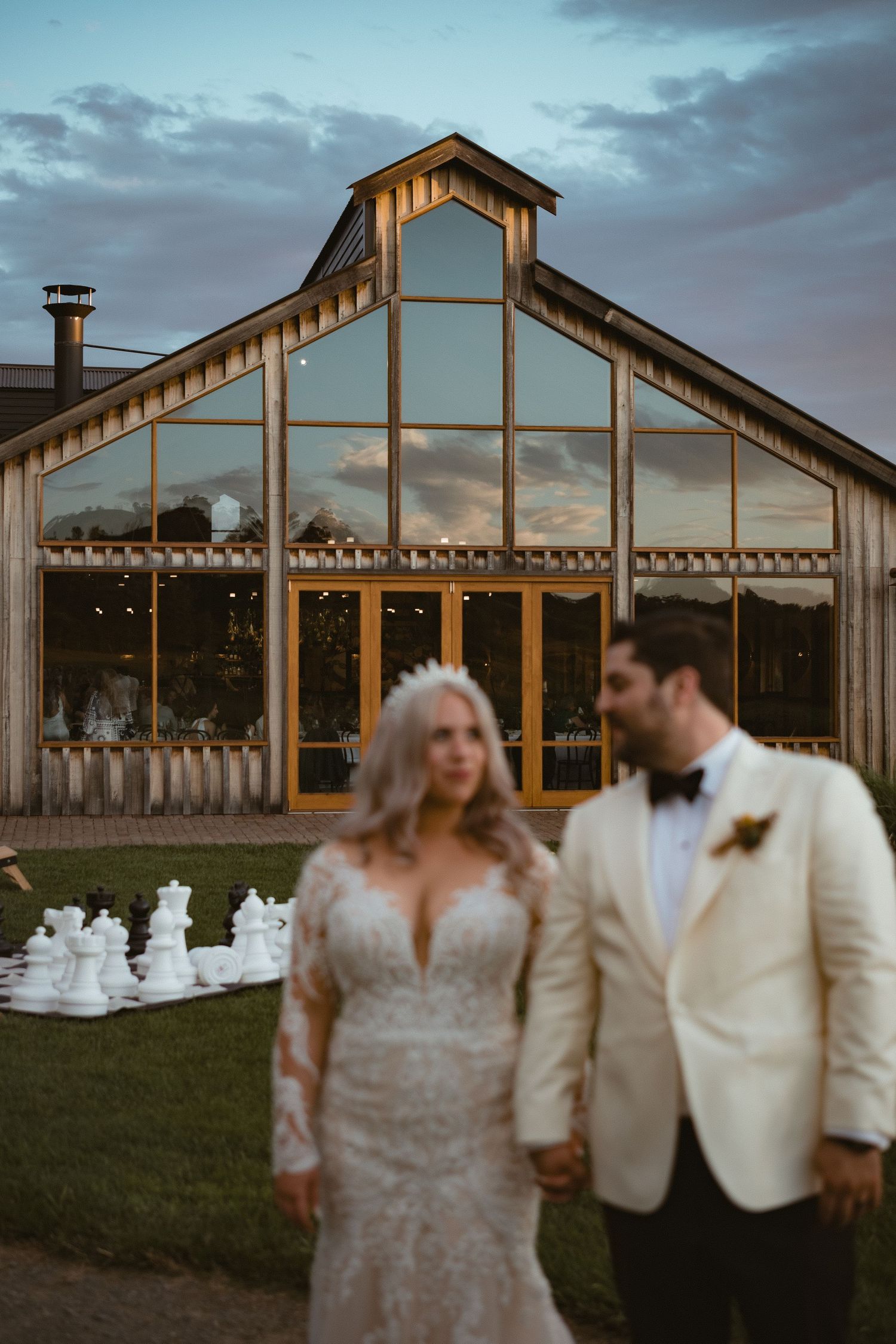 A modern glass wedding venue glows at dusk with string lights illuminating the exterior.