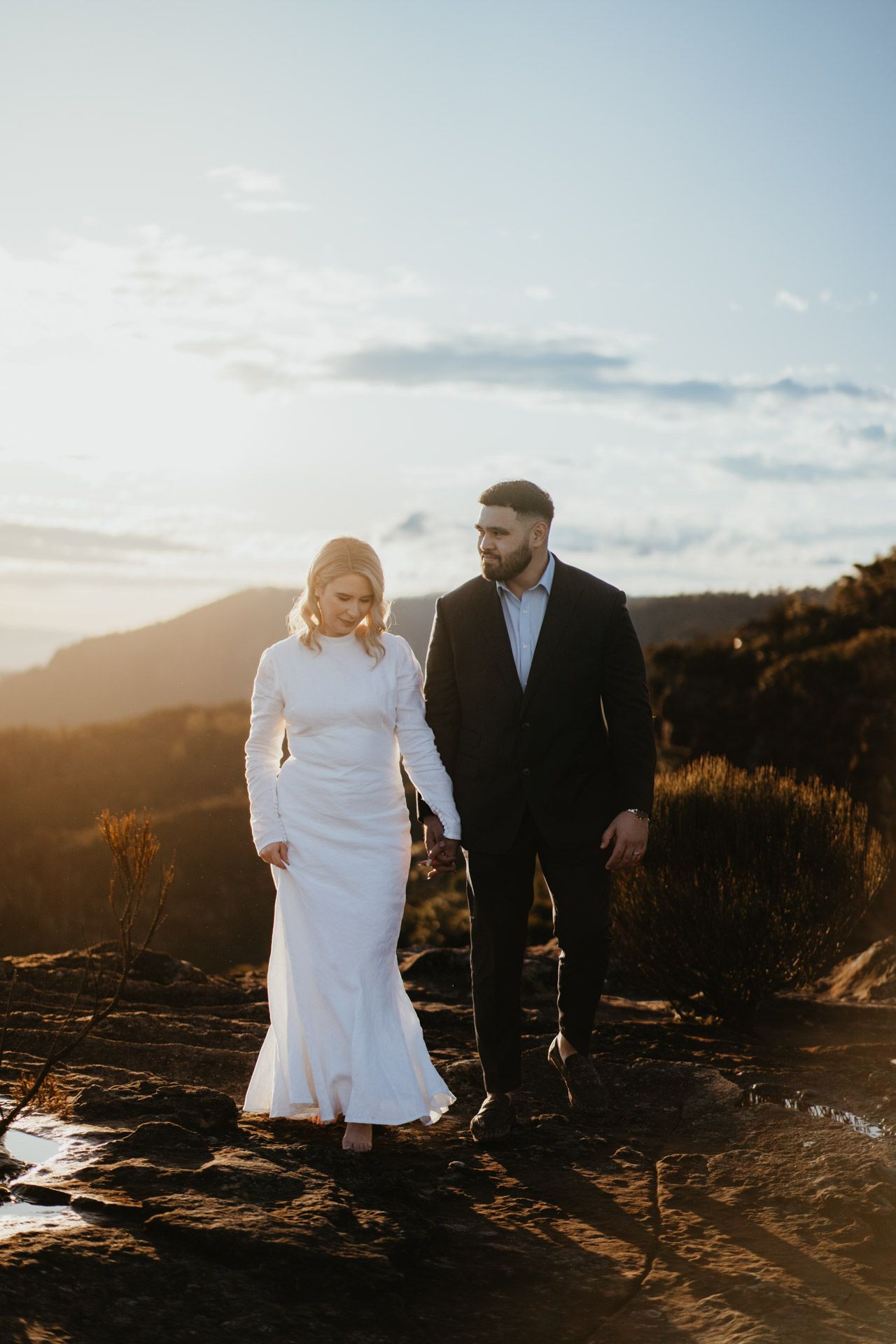 Couple in wedding attire walking along mountain ridge during golden hour.