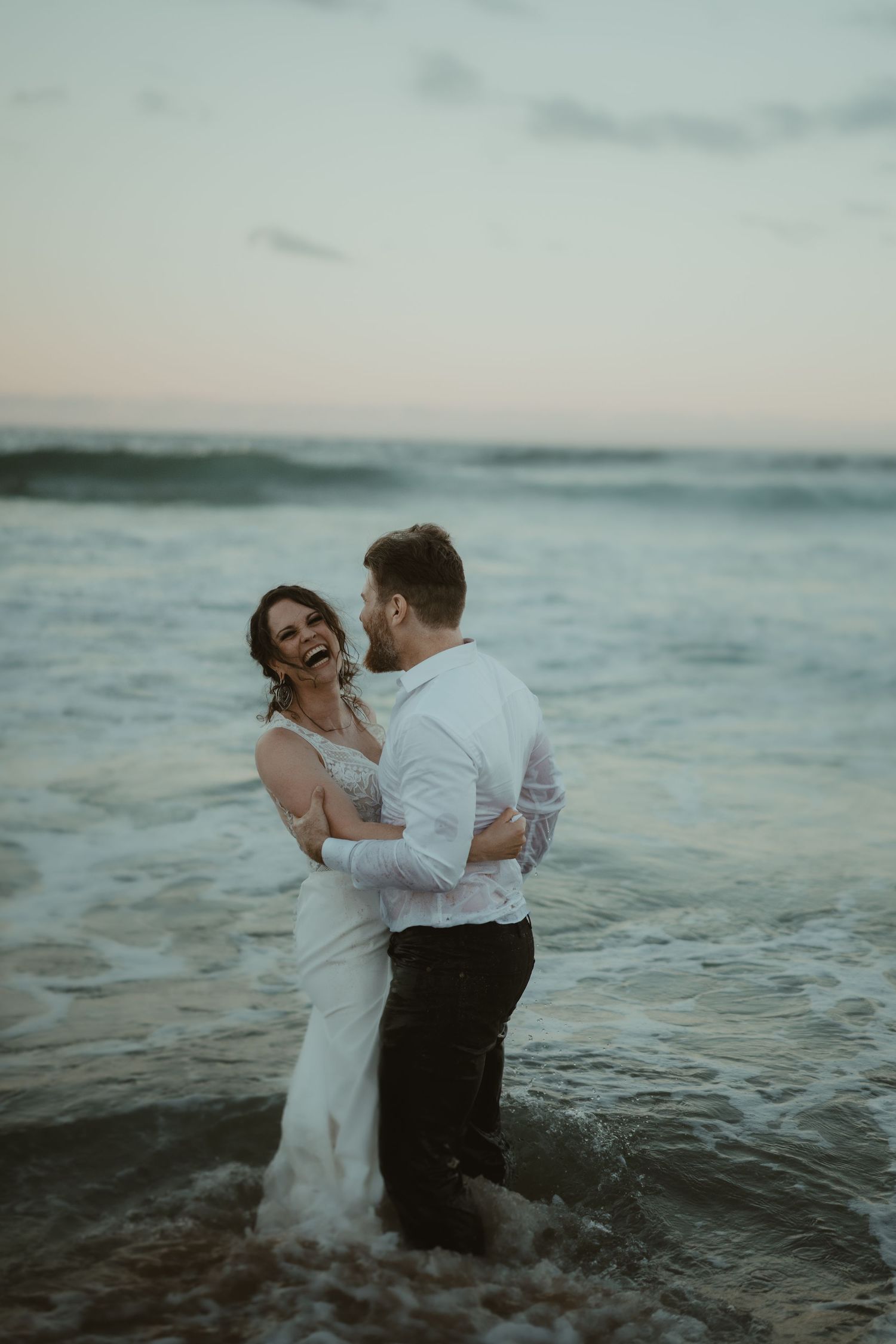 Couple in formal wear playing in ocean waves during sunset for romantic photoshoot.