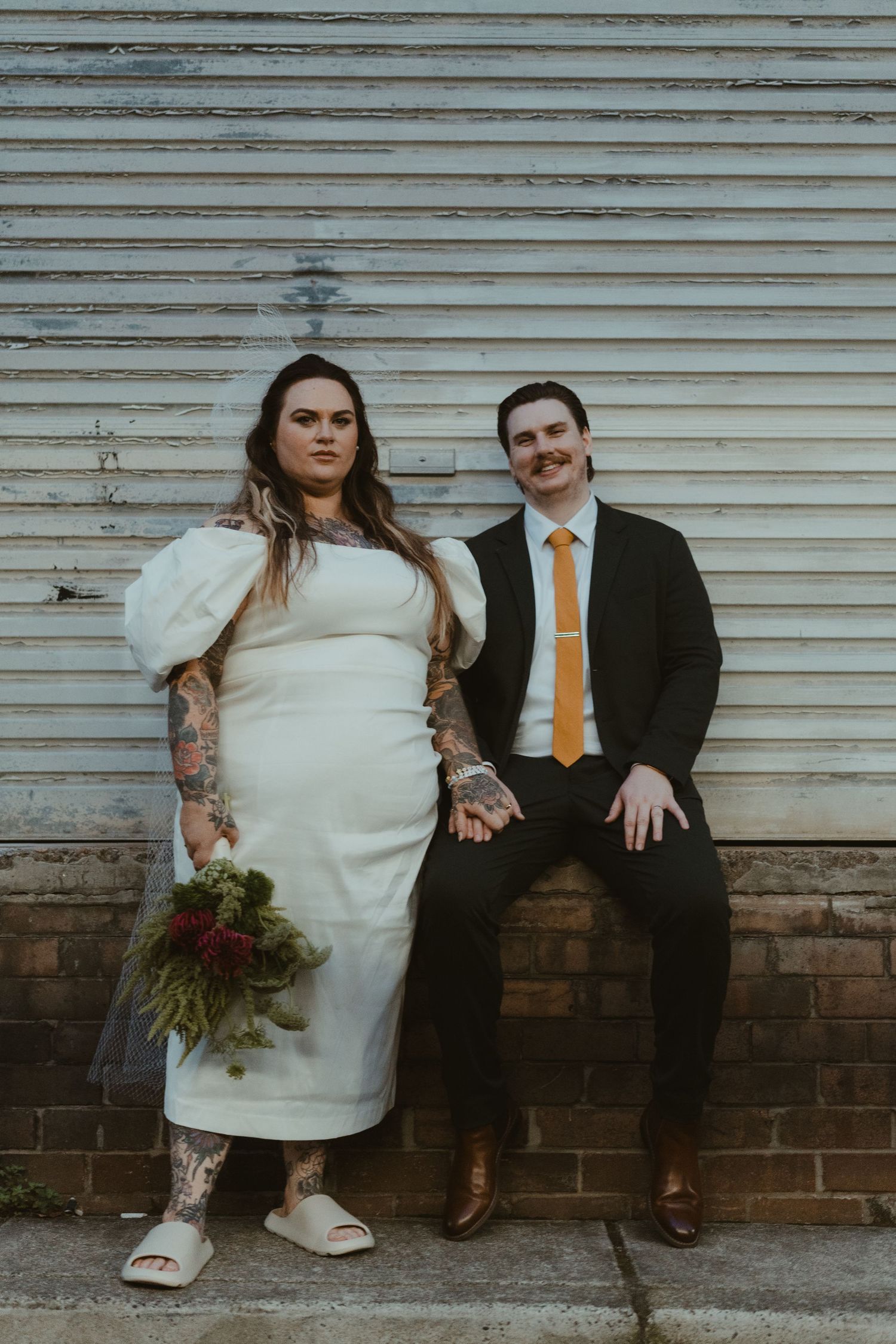 Couple in wedding attire sitting casually against a weathered wall with rustic charm.