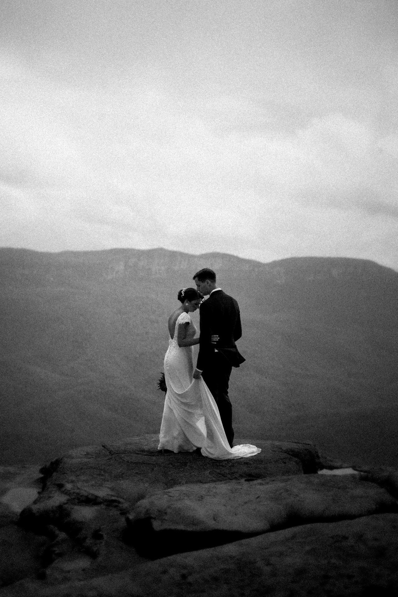Black and white wedding photography of a couple sitting on a mountainside overlooking a dramatic landscape.