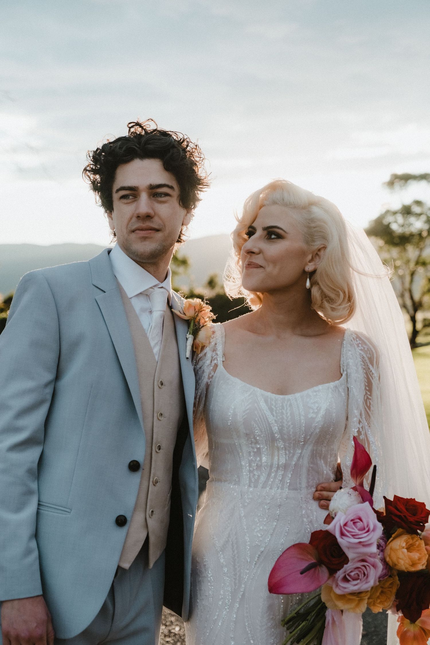 A couple in wedding attire poses together outdoors, with the bride holding a colorful bouquet of roses.