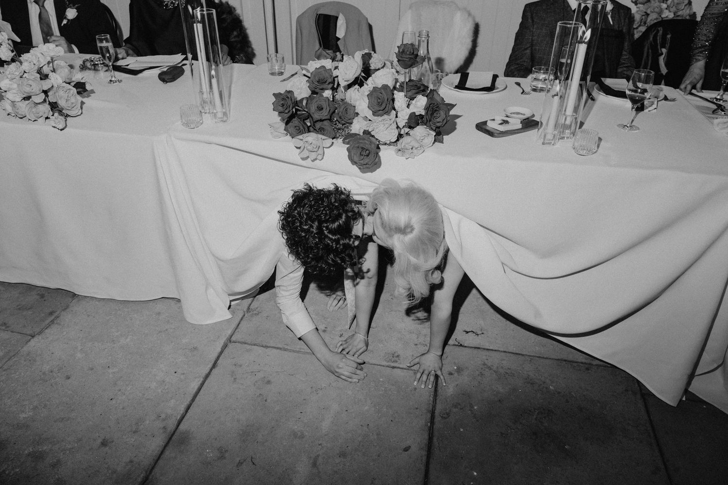 Couple together under a decorated wedding reception table with white linens.