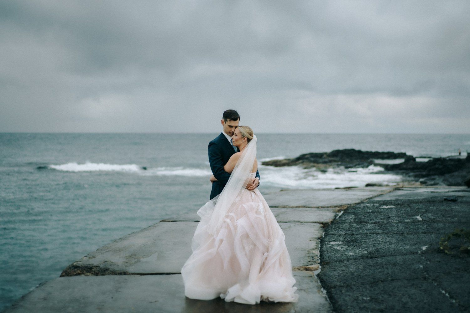 A dramatic coastal wedding photoshoot featuring a couple on black volcanic rocks against a moody ocean backdrop.