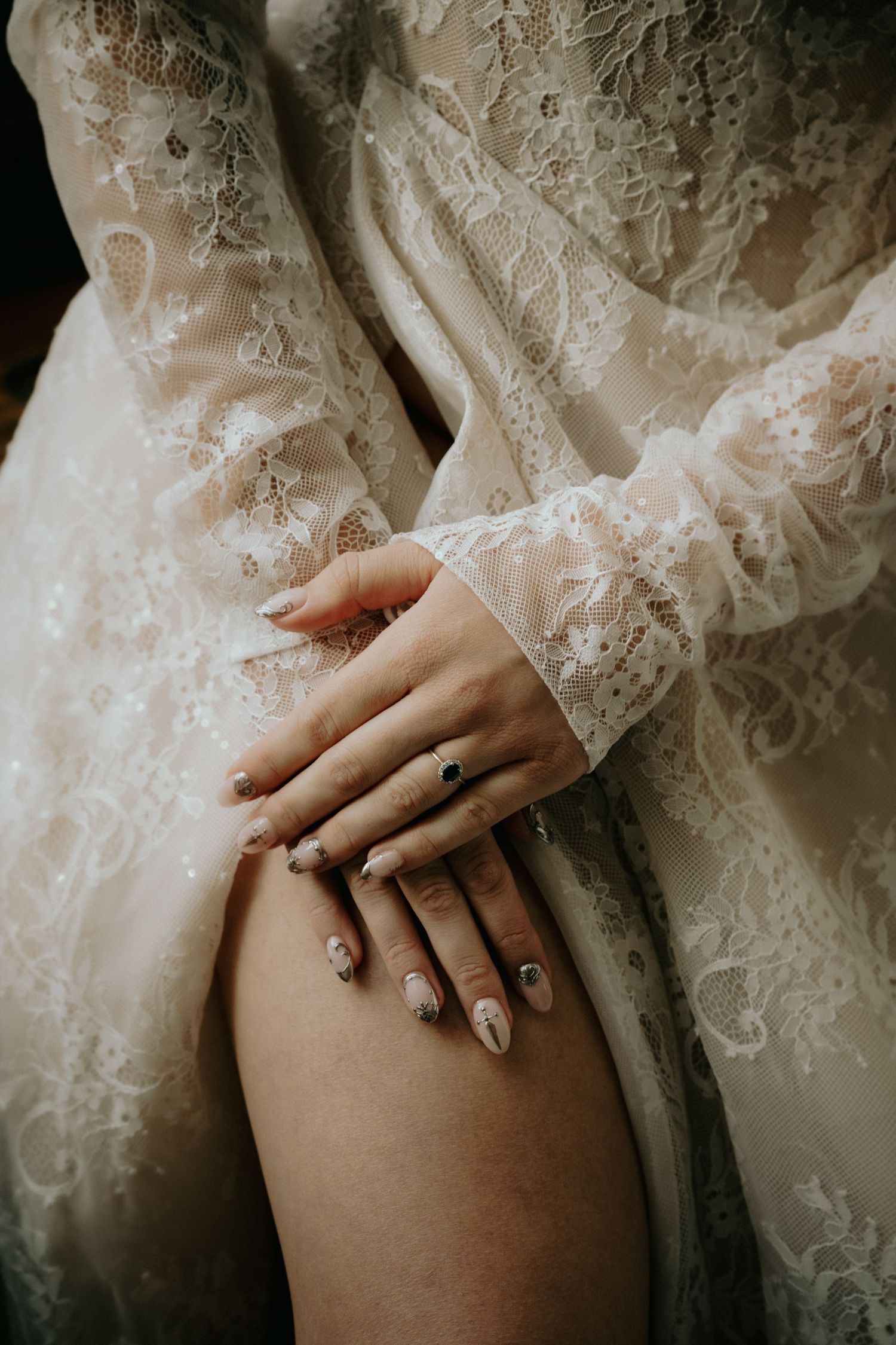 Close-up of a bride's hands with elegant nail art and a dark gemstone ring, resting on her leg in a lace bridal robe.