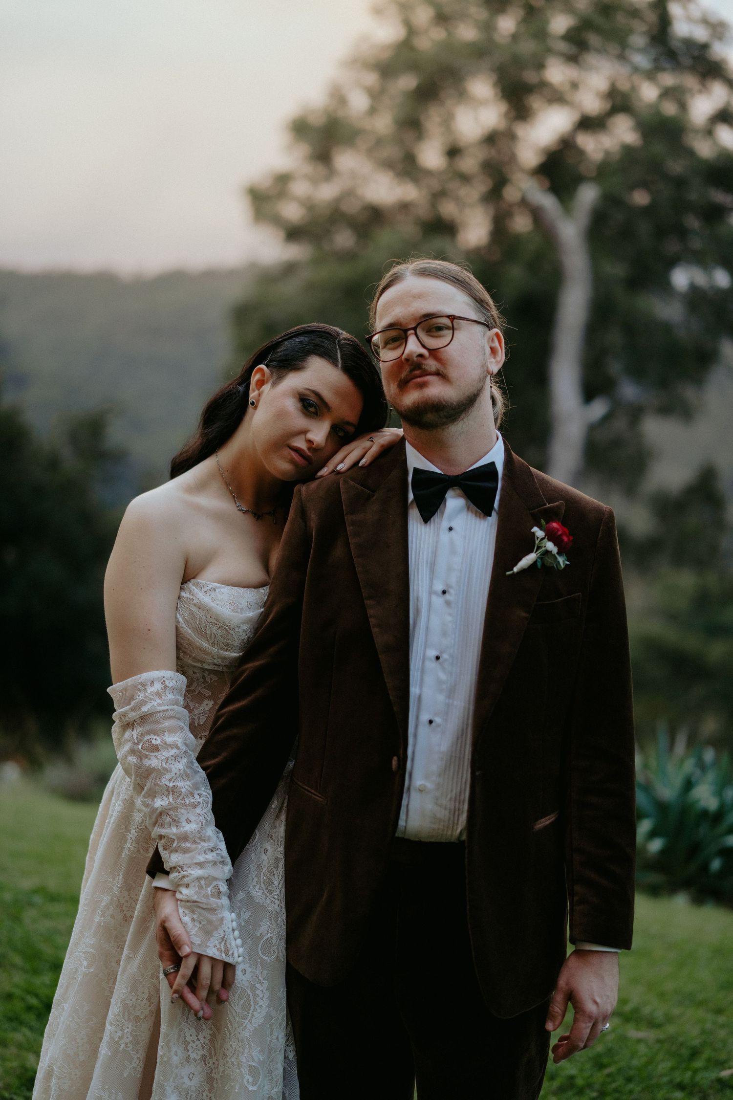A bride in a lace gown leans on a groom in a brown velvet tuxedo outdoors at golden hour.