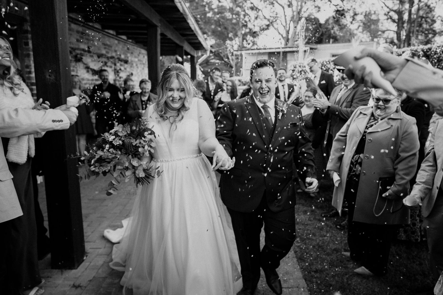 Wedding guests toss confetti over celebrating couple during outdoor ceremony exit.