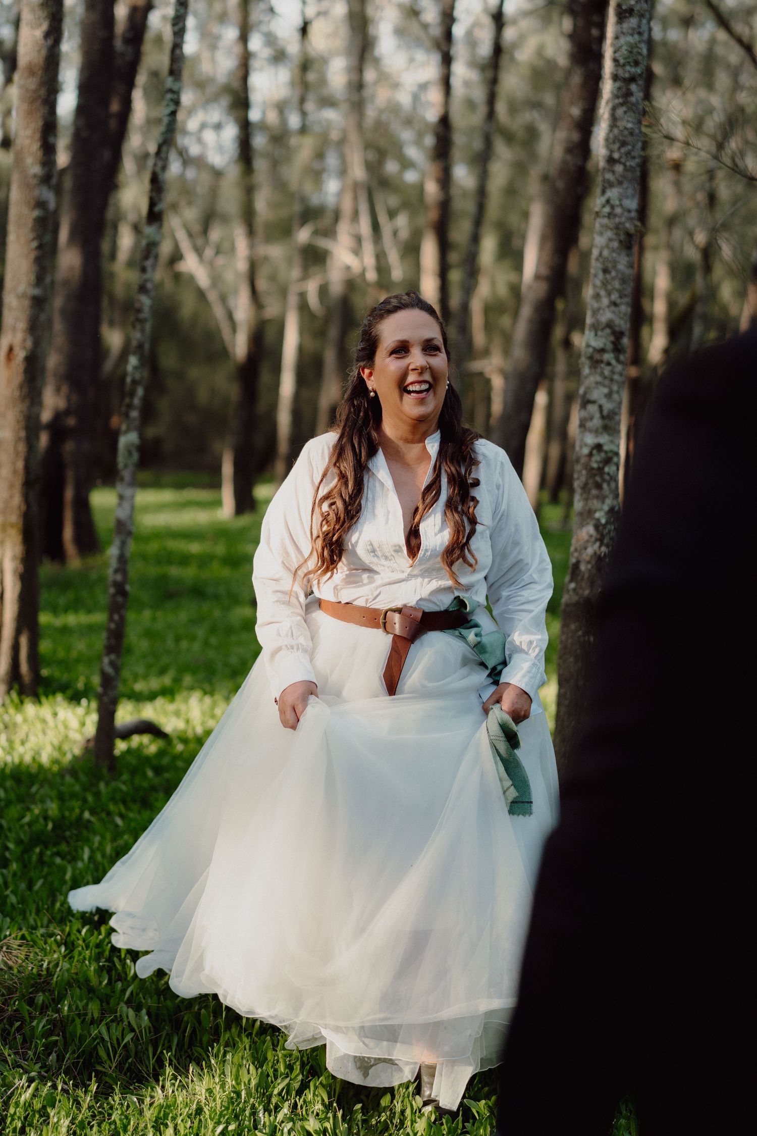 Person in white wedding dress laughs joyfully while sitting in a sunlit forest setting.