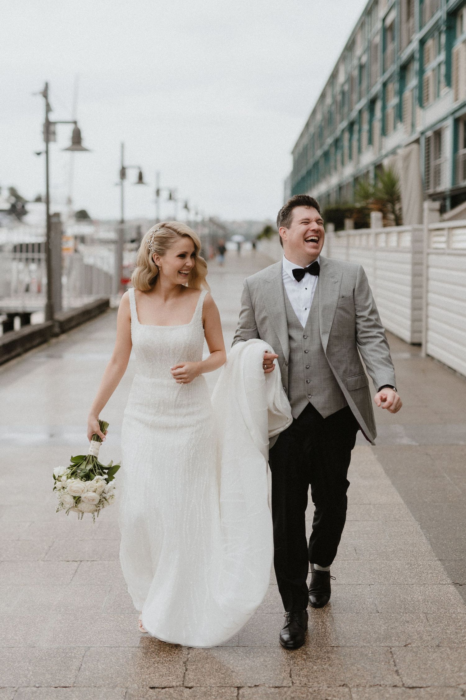 Joyful couple laughing while walking along an urban boardwalk on their wedding day.