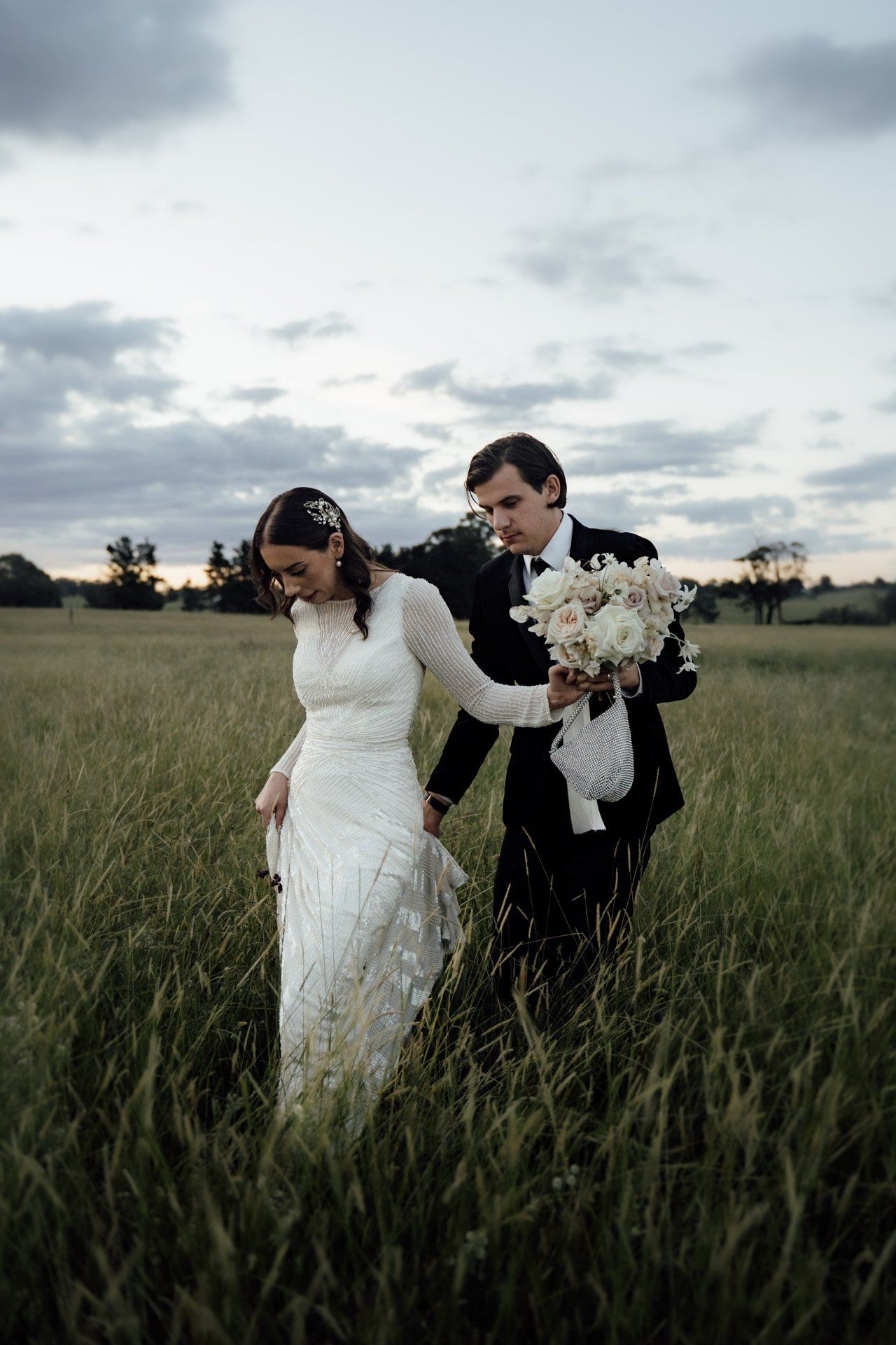Couple playfully runs through a tall grass field at sunset in formal wedding attire.