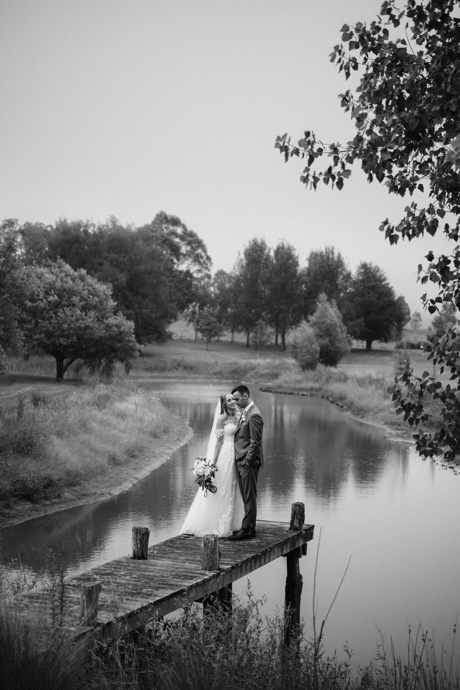 Black and white wedding portrait of a couple on a wooden dock by a serene lake.