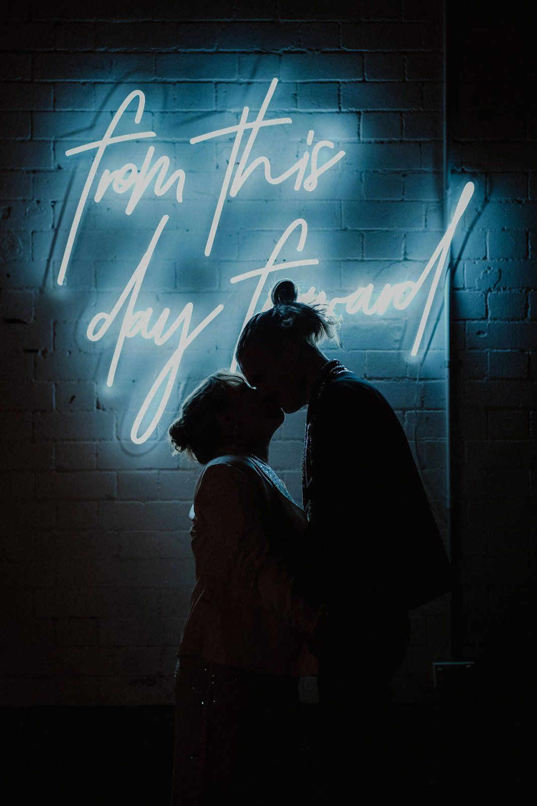 Silhouettes of a couple against a brick wall with blue neon lighting creating a moody atmosphere.