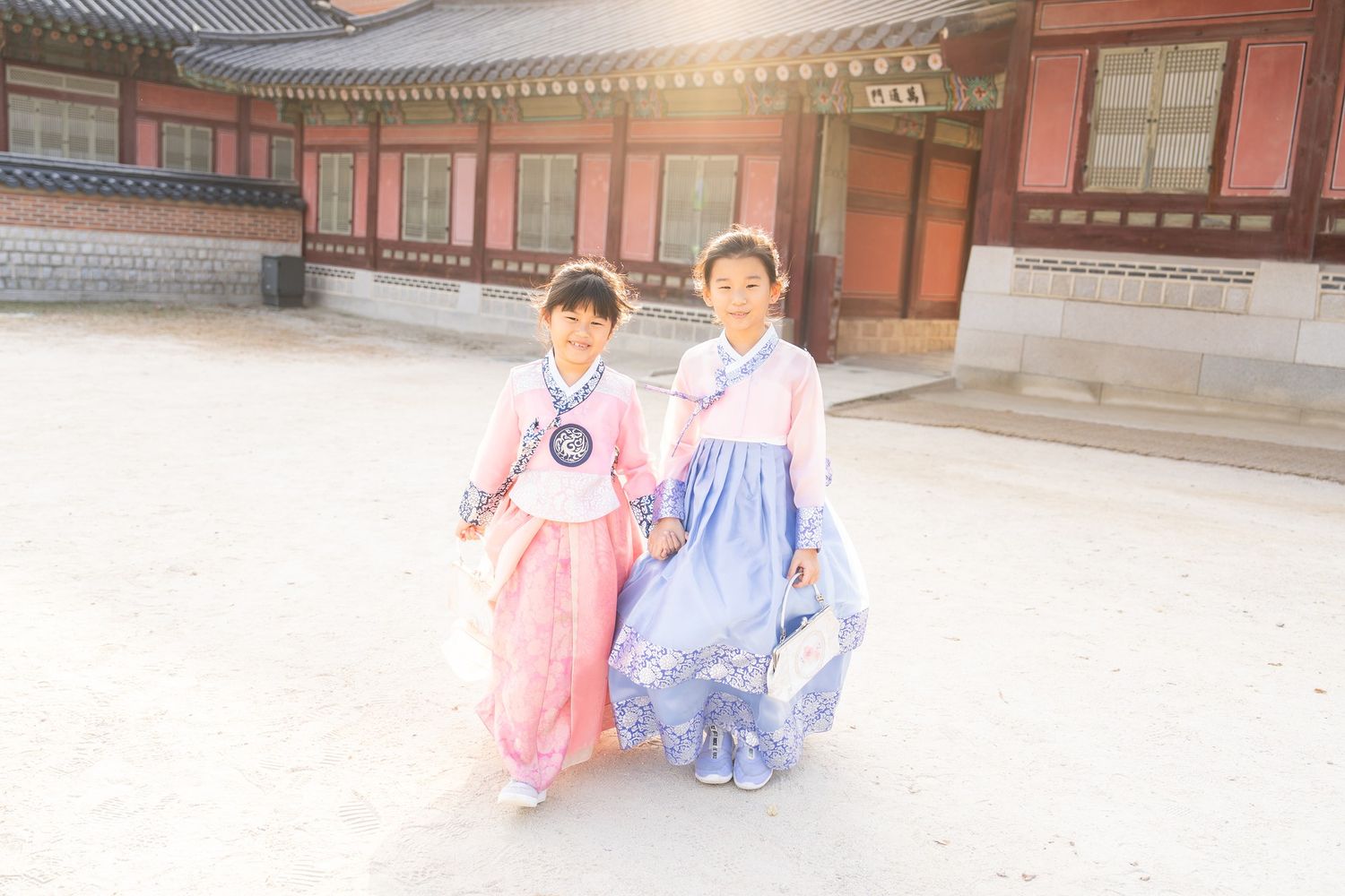 Two people wearing traditional Korean hanbok dresses pose in a courtyard of a historic palace in soft sunlight.