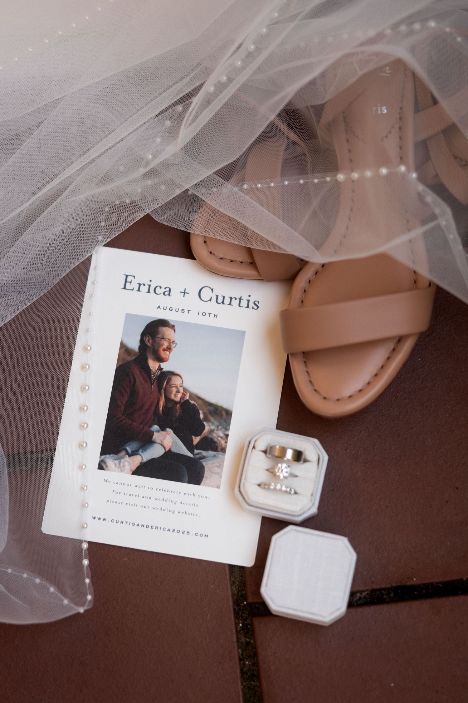 Wedding invitation and details card with veil and ring box on brown surface.