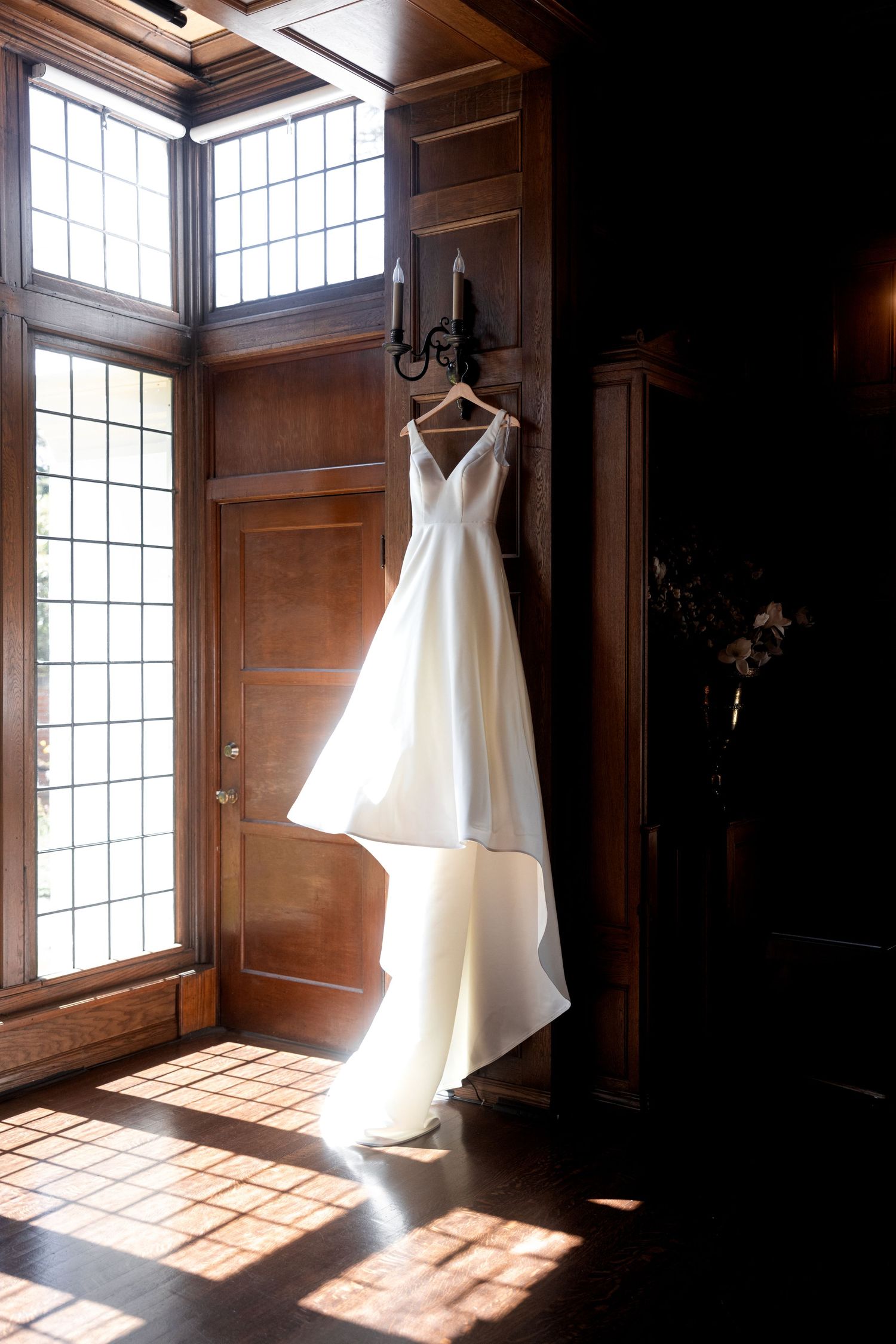 A white wedding dress hangs in a sunlit room with wooden walls and large windows creating dramatic lighting.