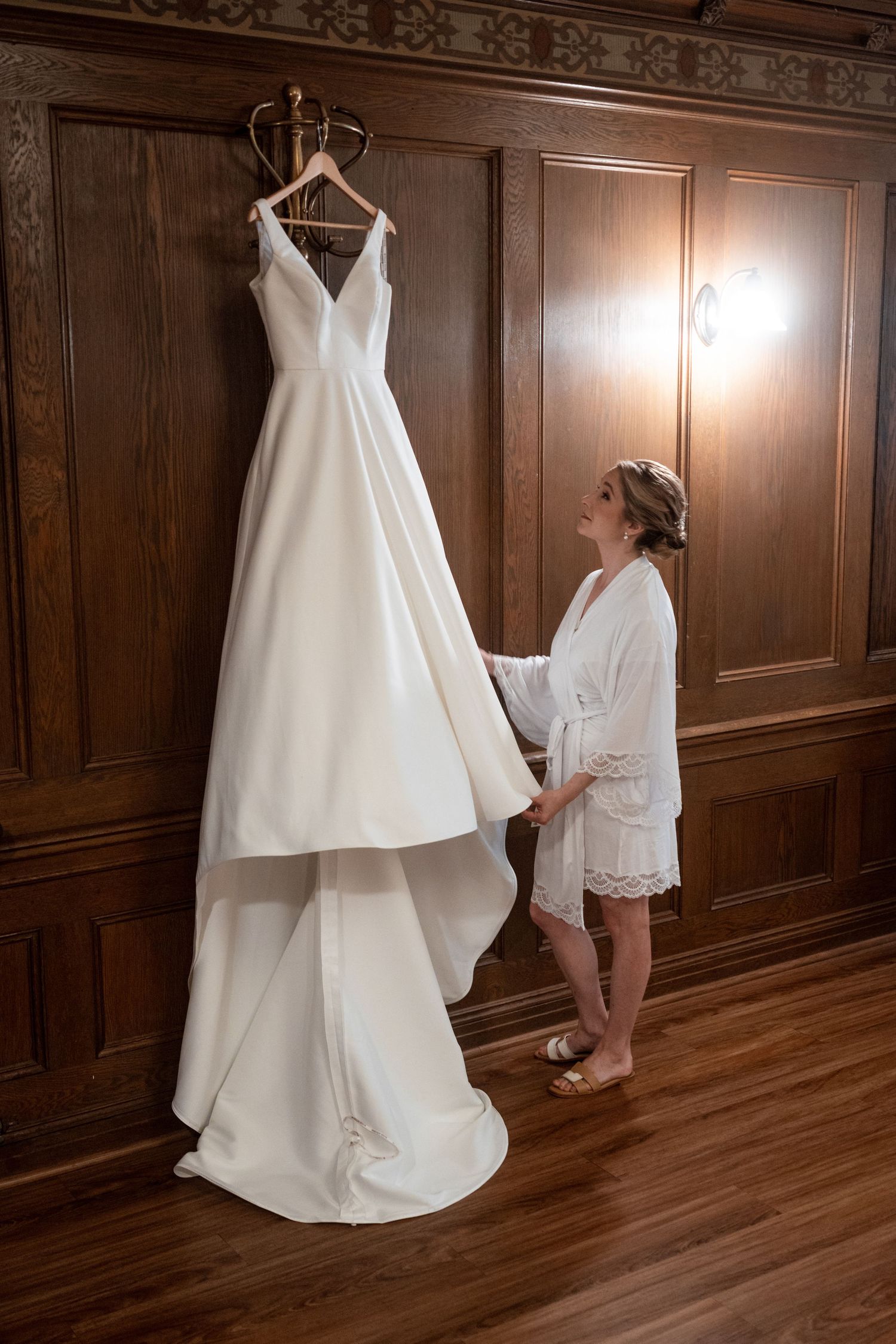 A white wedding dress hanging in a wood-paneled room during bridal preparations.