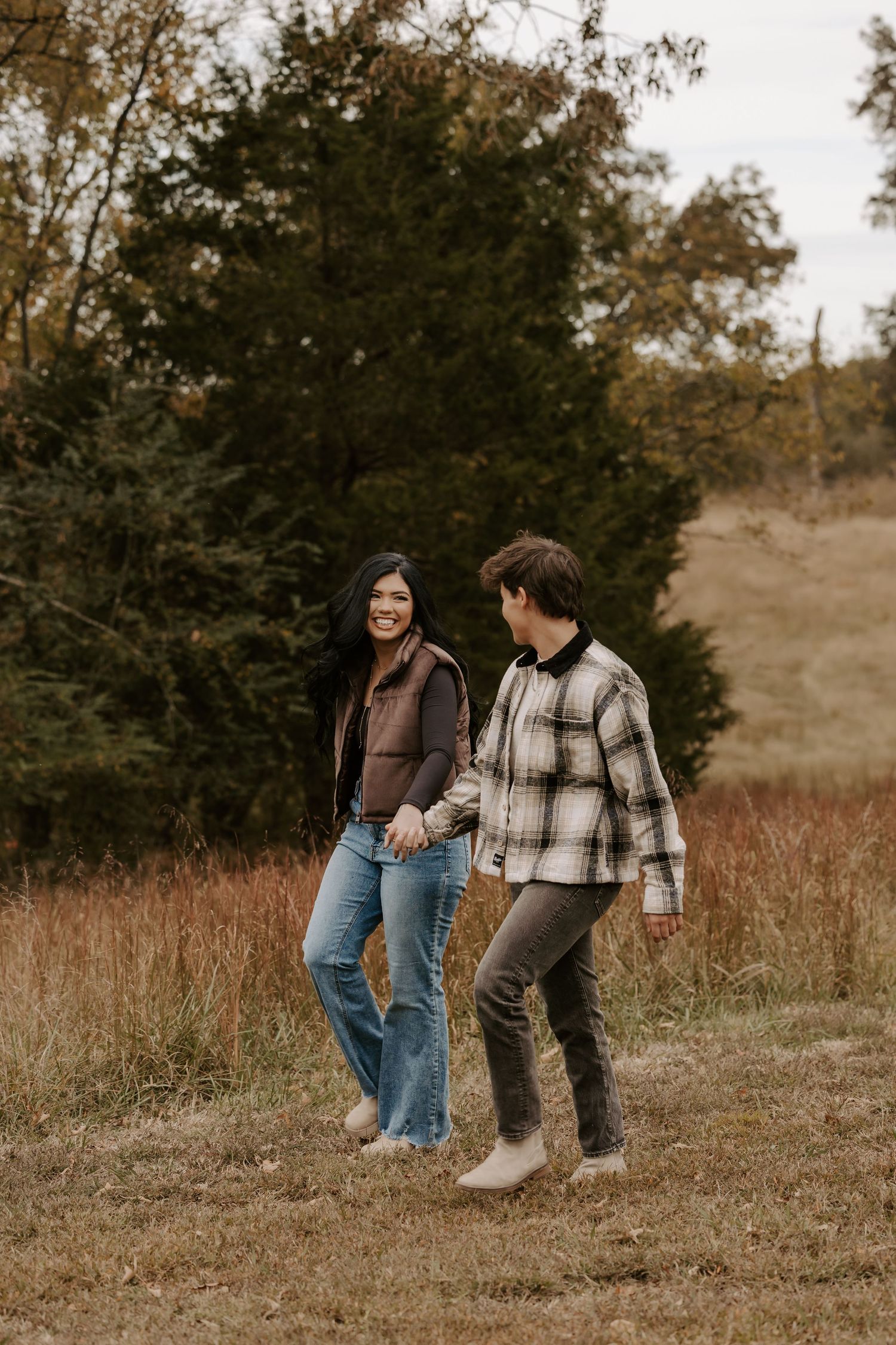 Couple walking together through a rustic field with fall foliage in the background.