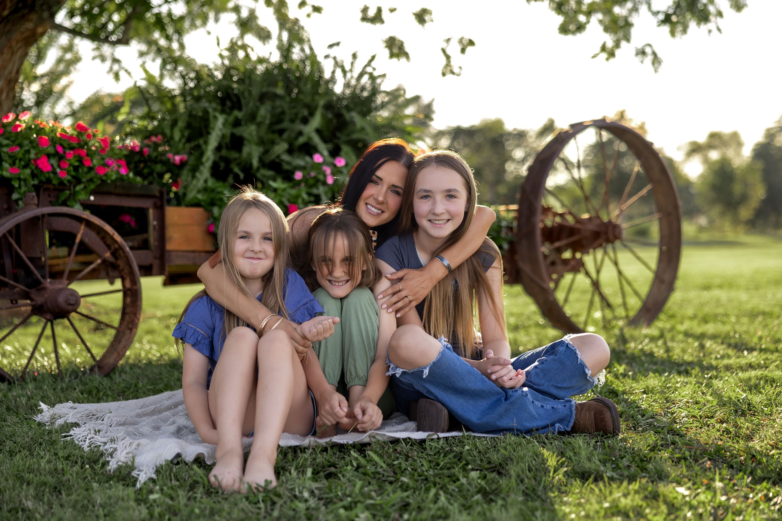 Casual outdoor family portrait with people lounging on blanket near rustic wagon wheel.