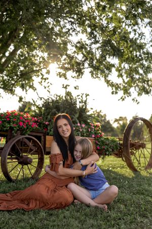 Intimate moment shared on grass near antique wagon with flowering plants in golden evening light.