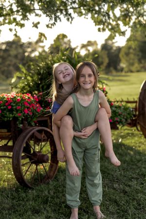 Piggyback ride moment captured in a garden setting with vintage wagon wheels and summer flowers.