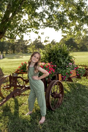 Casual pose against rustic garden decor with wagon wheels and colorful flower displays.